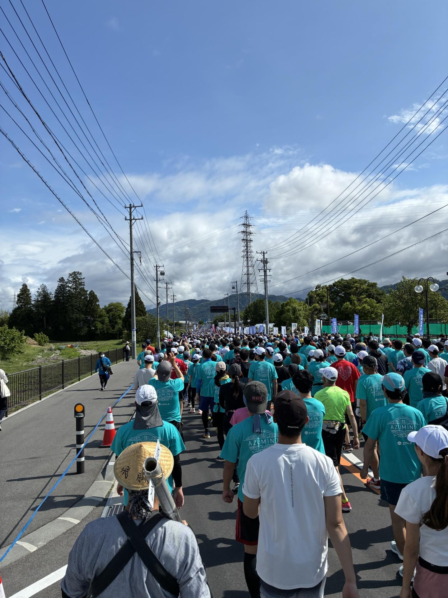 スタート直後の集団。水色のTシャツを着たランナーが道路いっぱいに広がって走っている。奥には雲のかかった山並み