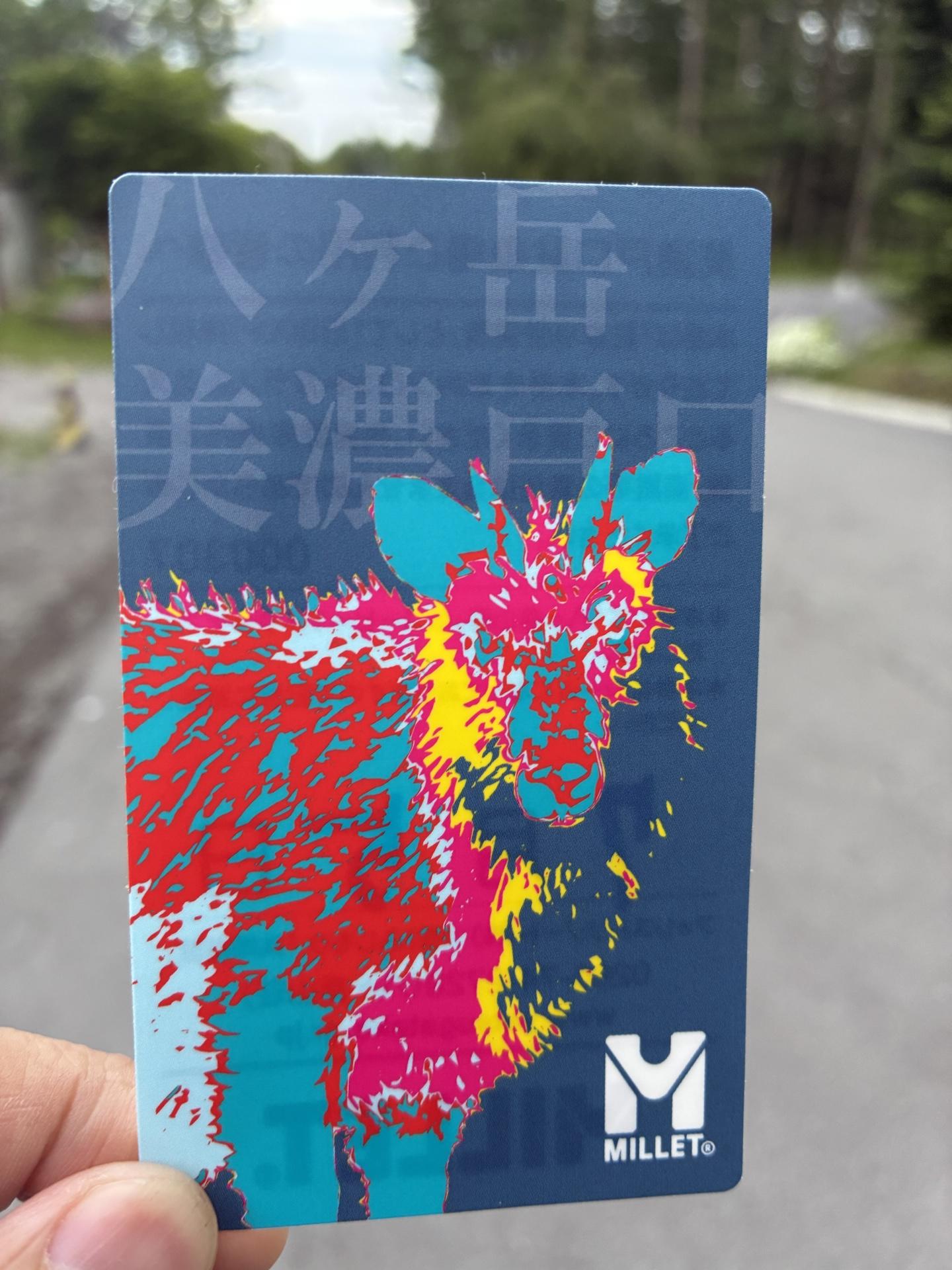 手に持った八ヶ岳美濃戸口の駐車券。カラフルな牛のイラストが印刷されたデザインで、ミレーのロゴが入っている
