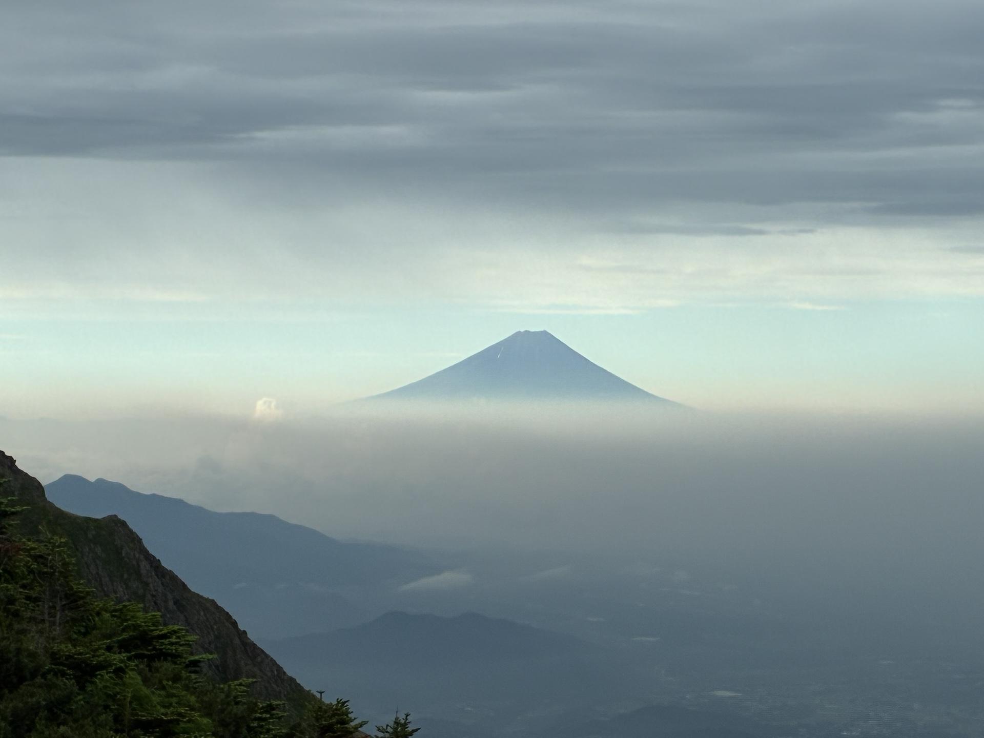雲海の上に浮かぶ富士山のシルエット。手前は霞んだ山並みで、空は薄曇り