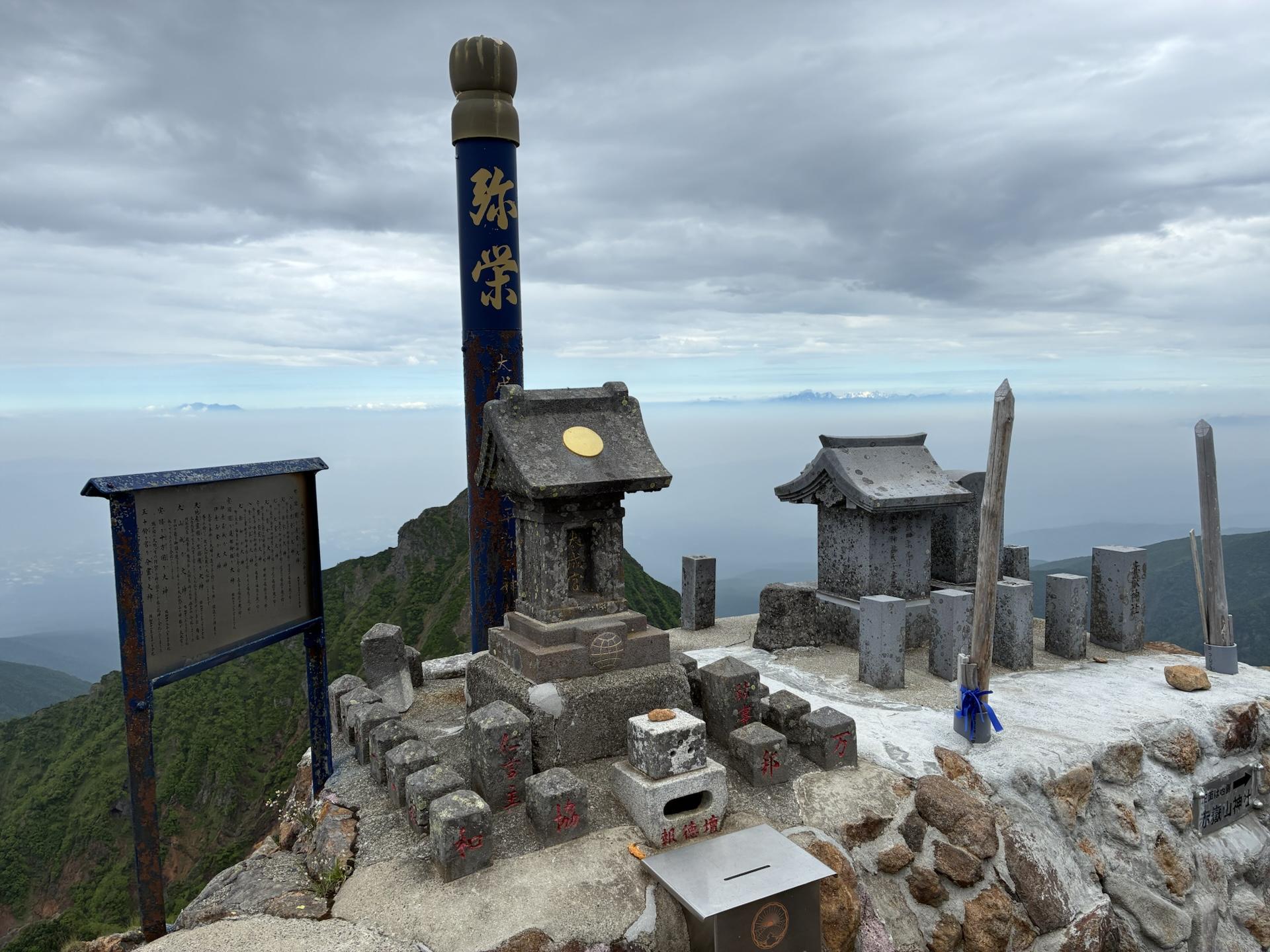 赤岳山頂に並ぶ二つの石造りの祠。周囲は岩に囲まれ、奥には雲の広がる空と遠い山並みが見える
