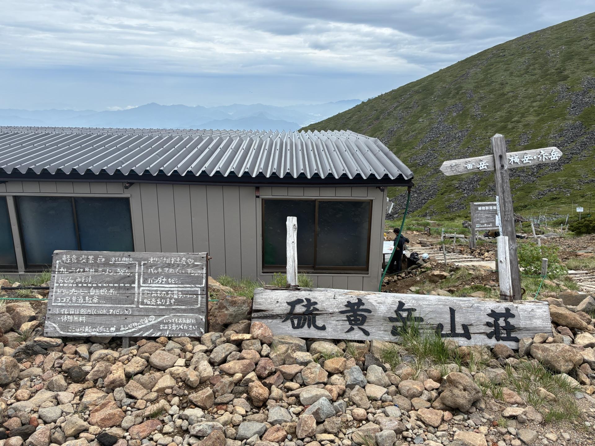 稜線の鞍部に建つ硫黄岳山荘。灰色の屋根の建物の前に道標が立ち、背後には緑の斜面が広がっている