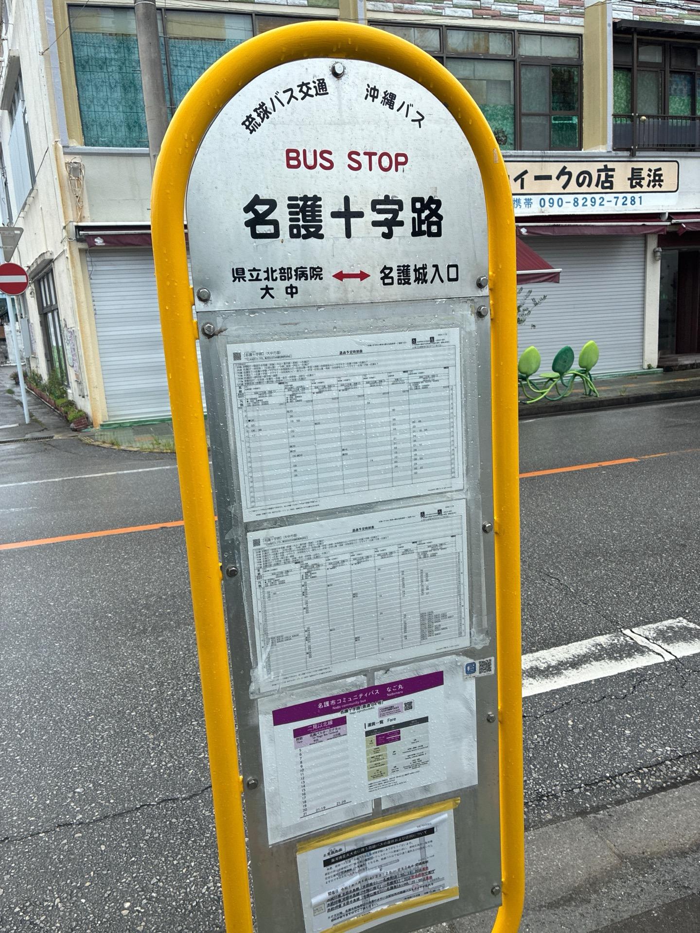 「名護十字路」と書かれた黄色いバス停の標識。後ろには沖縄の街並みと商店が並んでいる