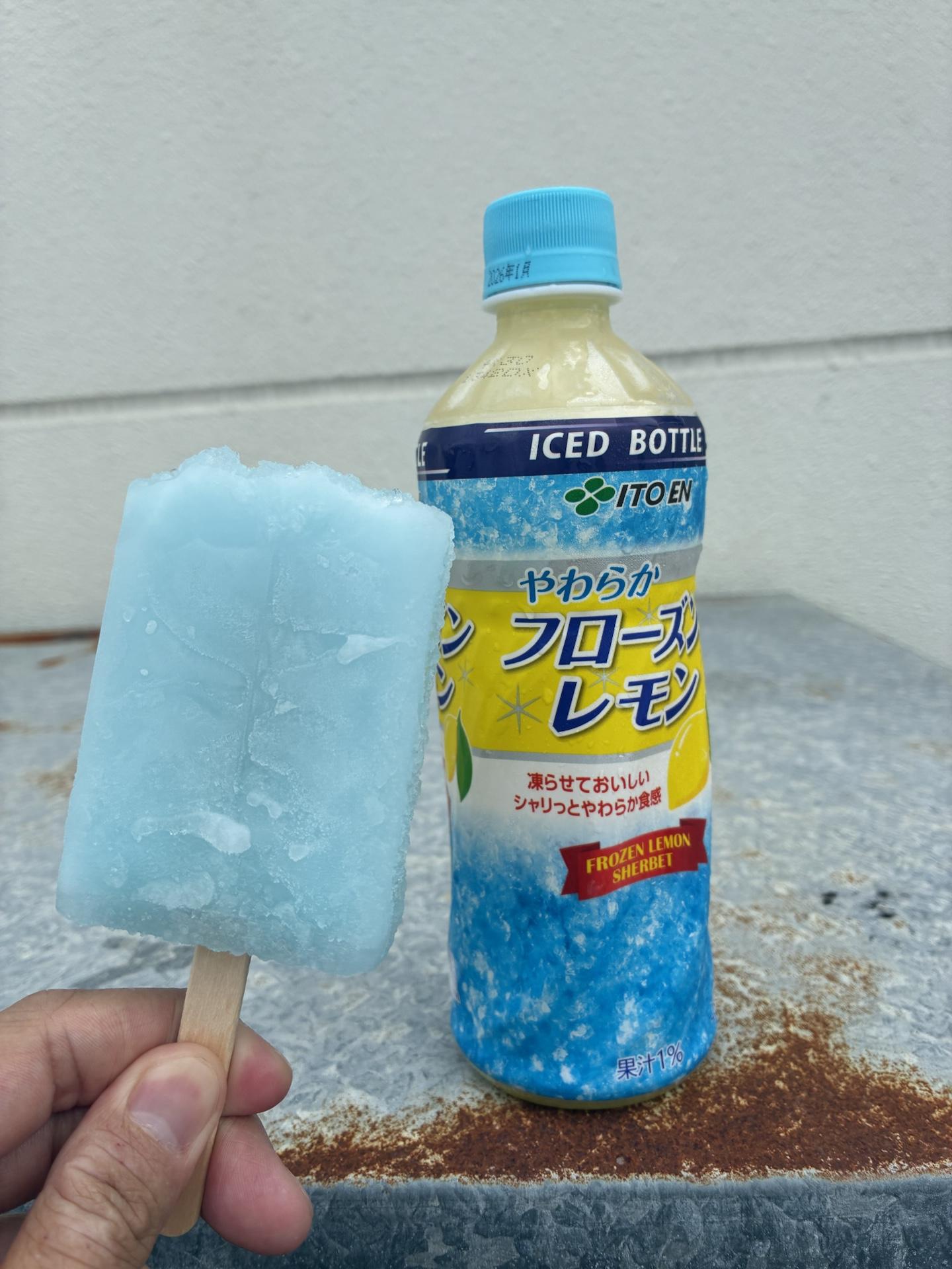 手に持った水色のかき氷バーと、レモン味のペットボトル飲料。背景はコンビニの駐車場のアスファルト