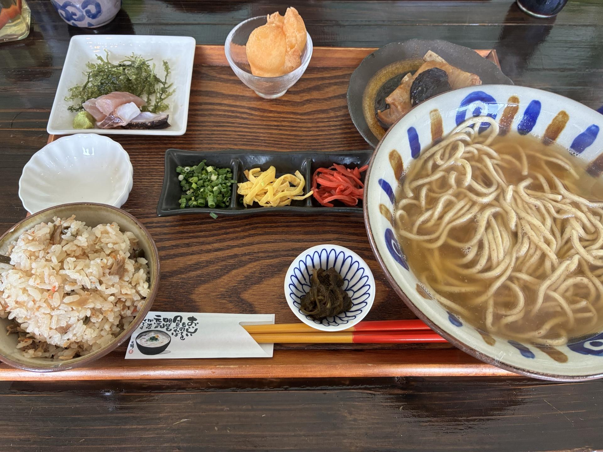 木の盆に並んだ沖縄そばの定食。太めの麺のそばに、ジューシー、小鉢、もずくなどが添えられている