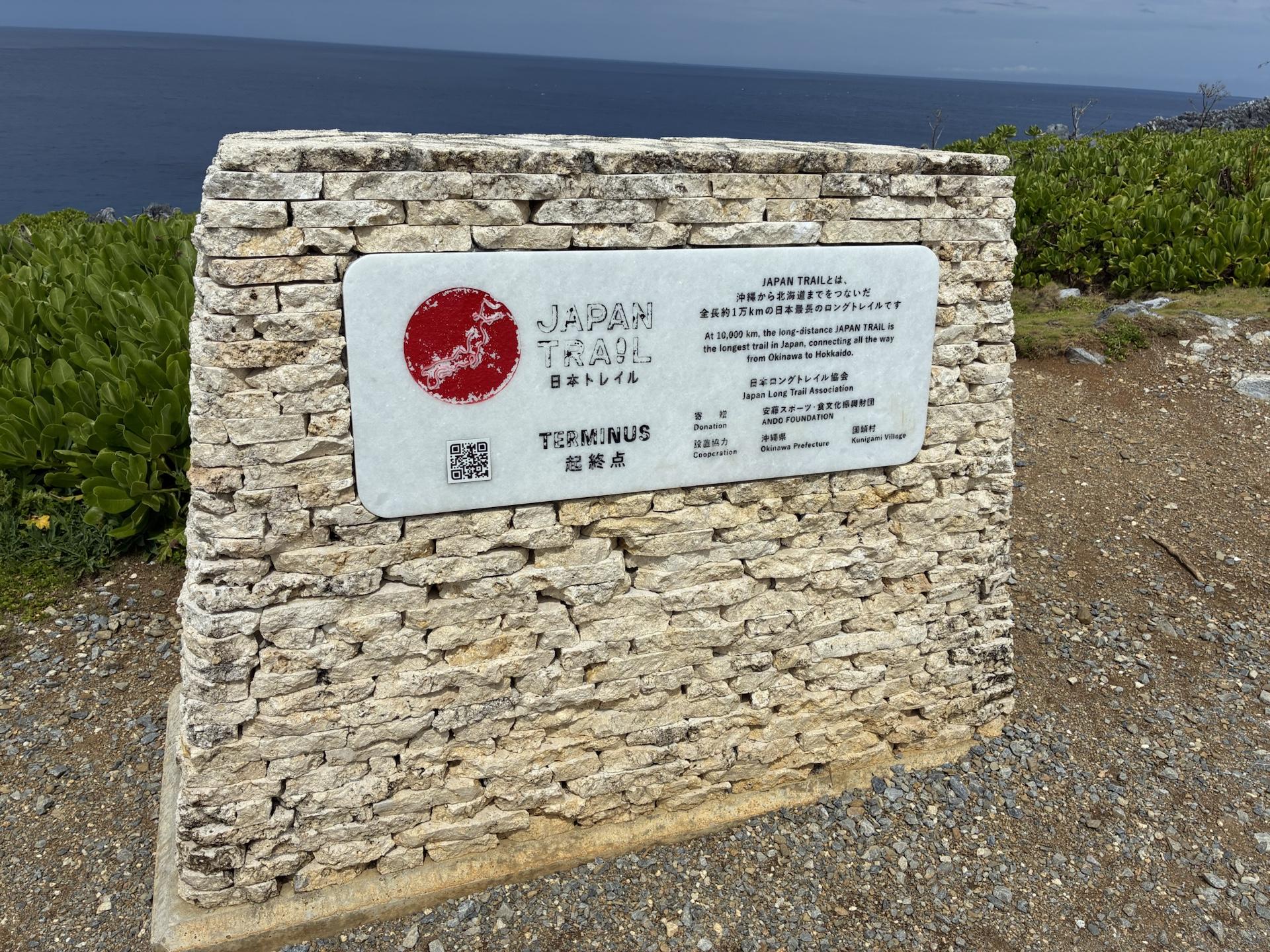 石を積んだ台に取り付けられた「JAPAN TRAIL TERMINUS 起終点」のプレート。背景は岬の草地と海