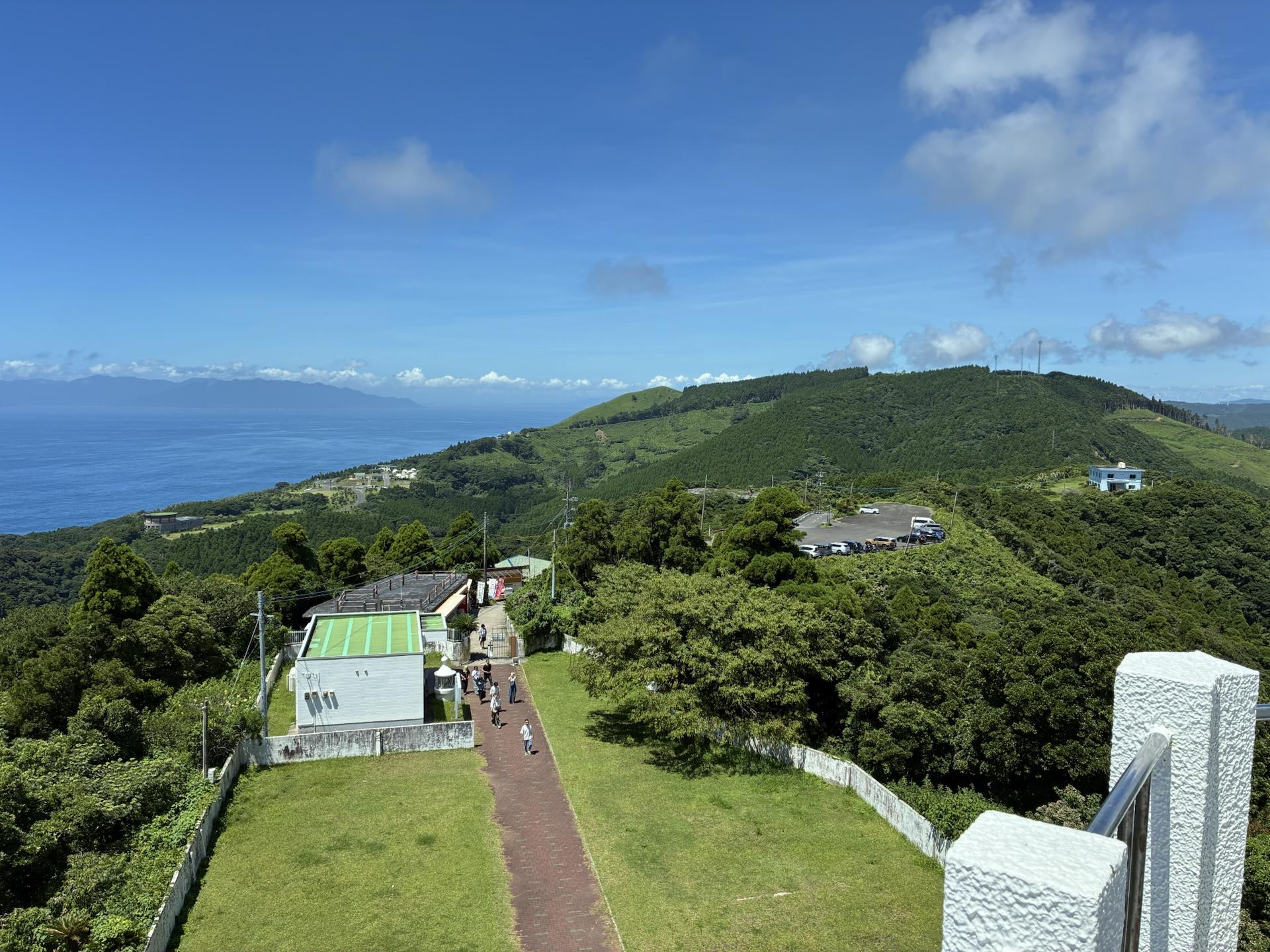 高台から見下ろした都井岬。緑におおわれた岬の斜面に白い建物と園路が続き、その先に青い海と対岸の山並みが広がっている