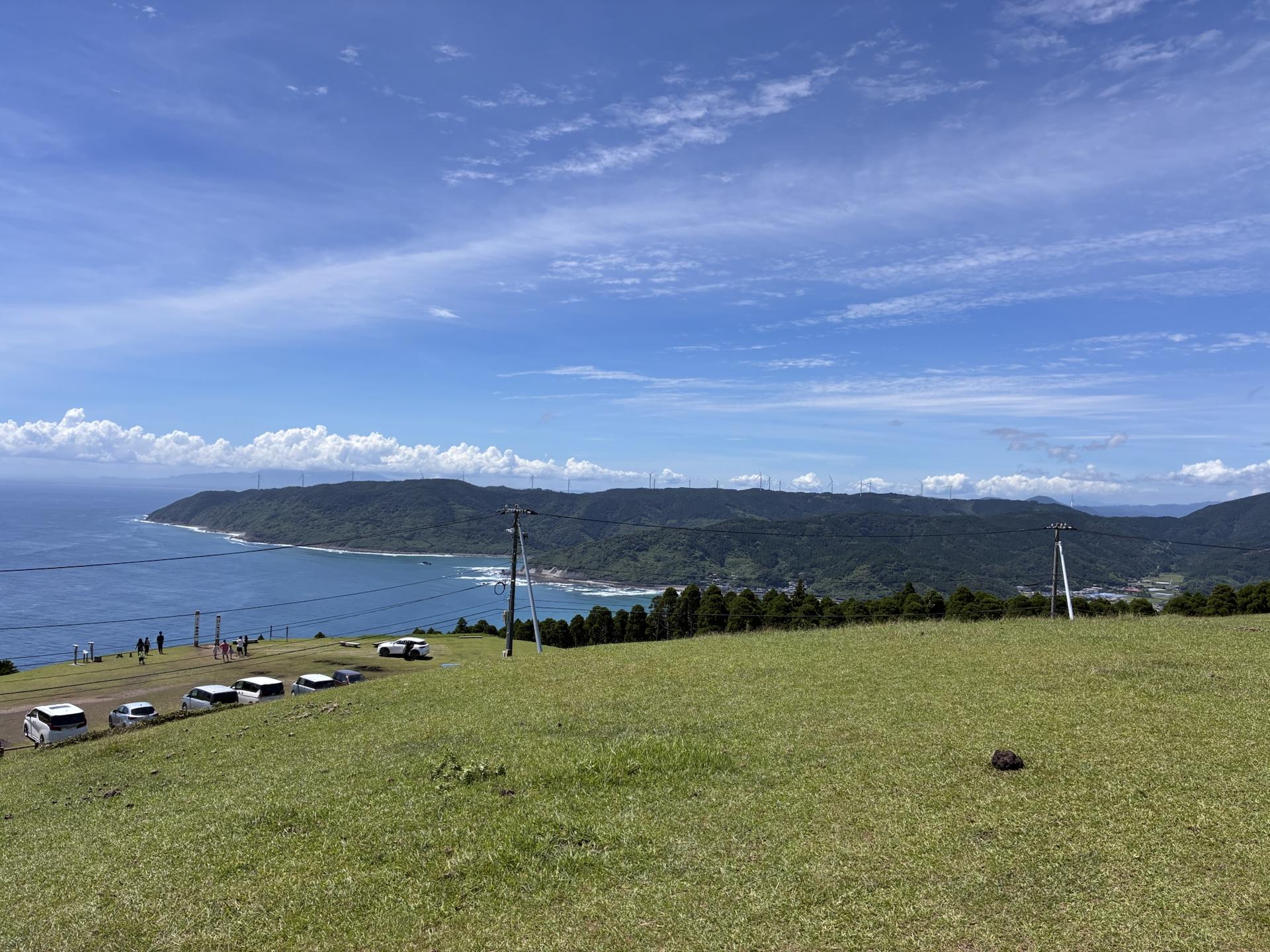 小松ヶ丘の広い草原。手前は刈られた草地で、その先に湾曲した海岸線と青い海、遠くに山並みが続いている。草原の端には駐車場に停まった車が並ぶ