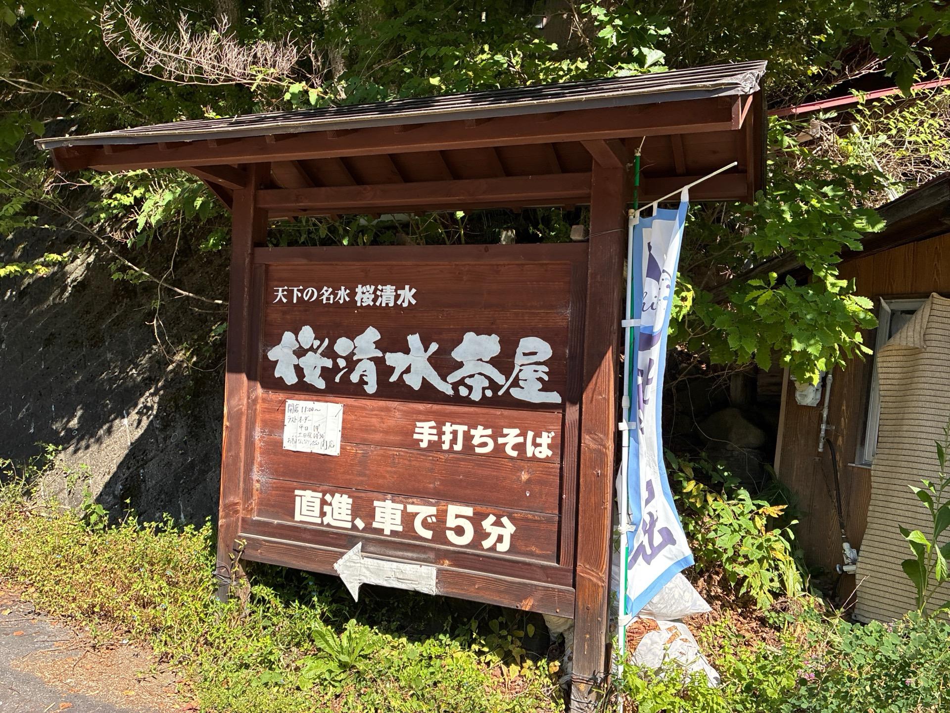 道路脇に立つ「天下の名水 桜清水 桜清水茶屋 手打ちそば 直進、車で5分」と書かれた木製の大きな看板。屋根付きの立派なつくり