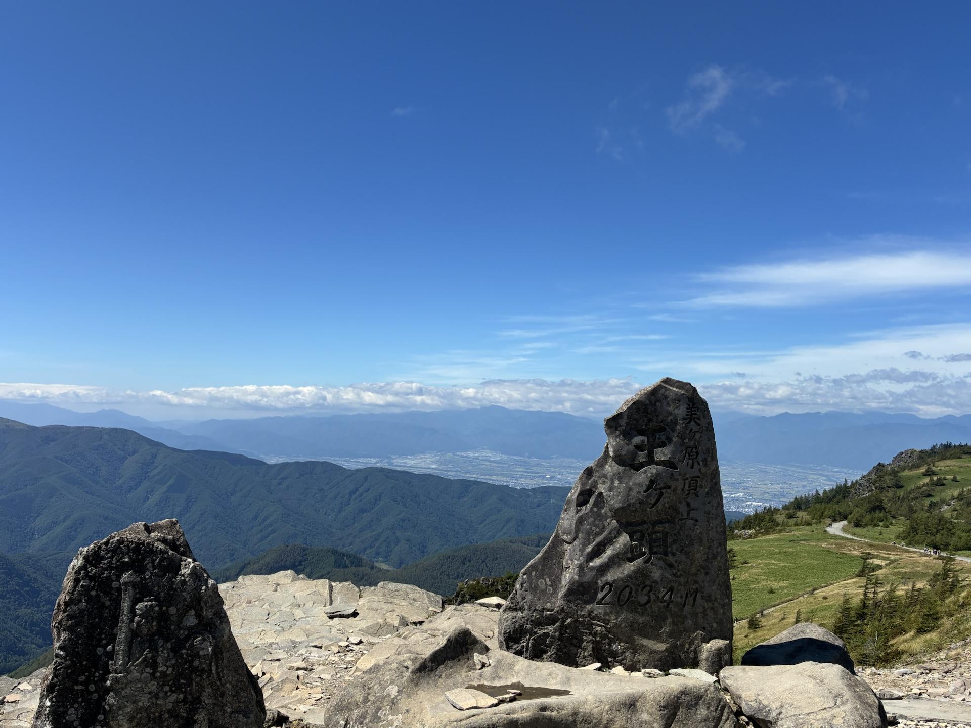 王ヶ頭の山頂に立つ大きな岩。手前はザレた地面で、奥には雲の上に連なる遠くの山並みが霞んで見える