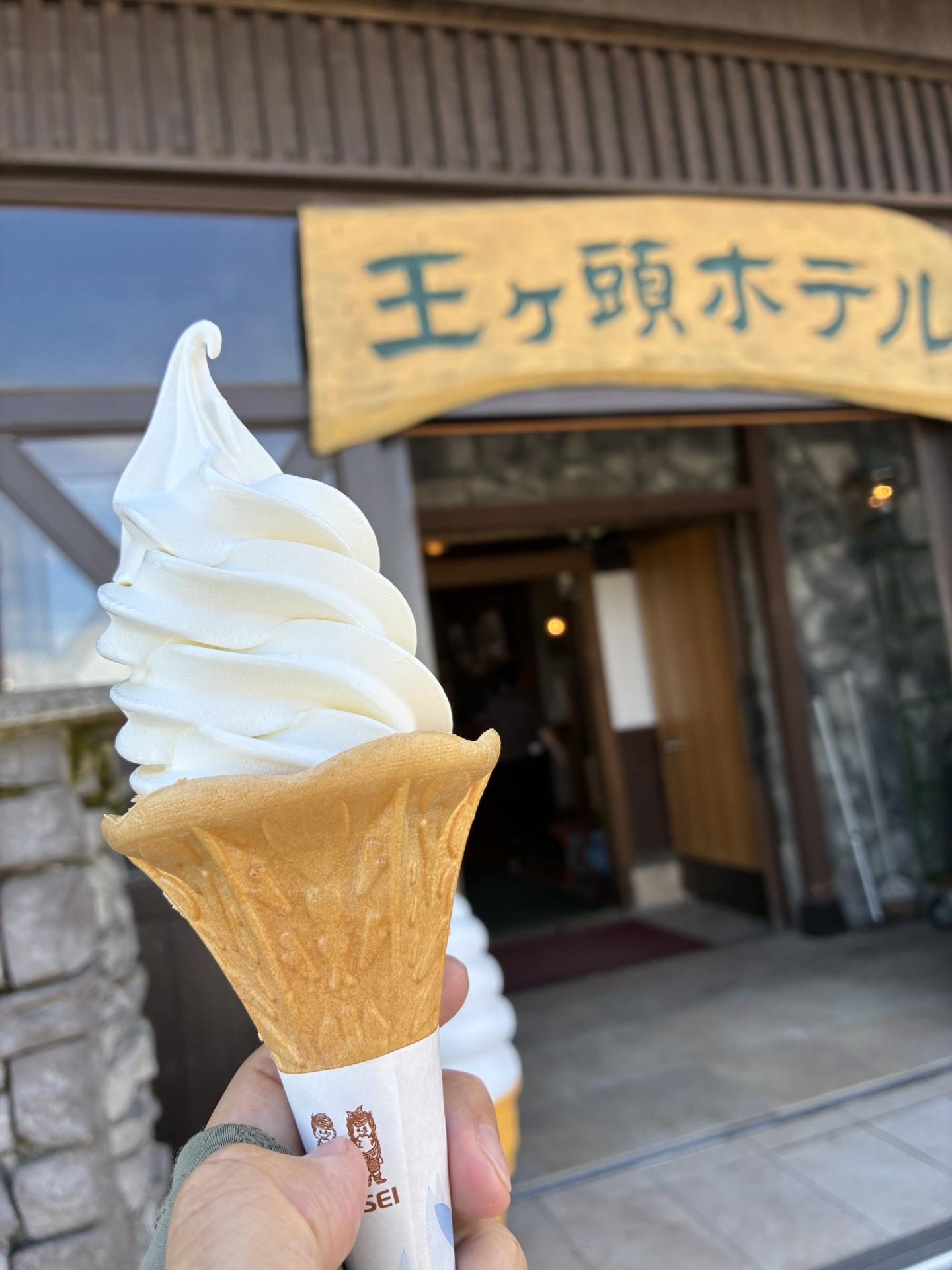 王ヶ頭ホテルの入口前で手に持ったソフトクリーム。背景に「王ヶ頭ホテル」と書かれた看板と木造の建物が写っている