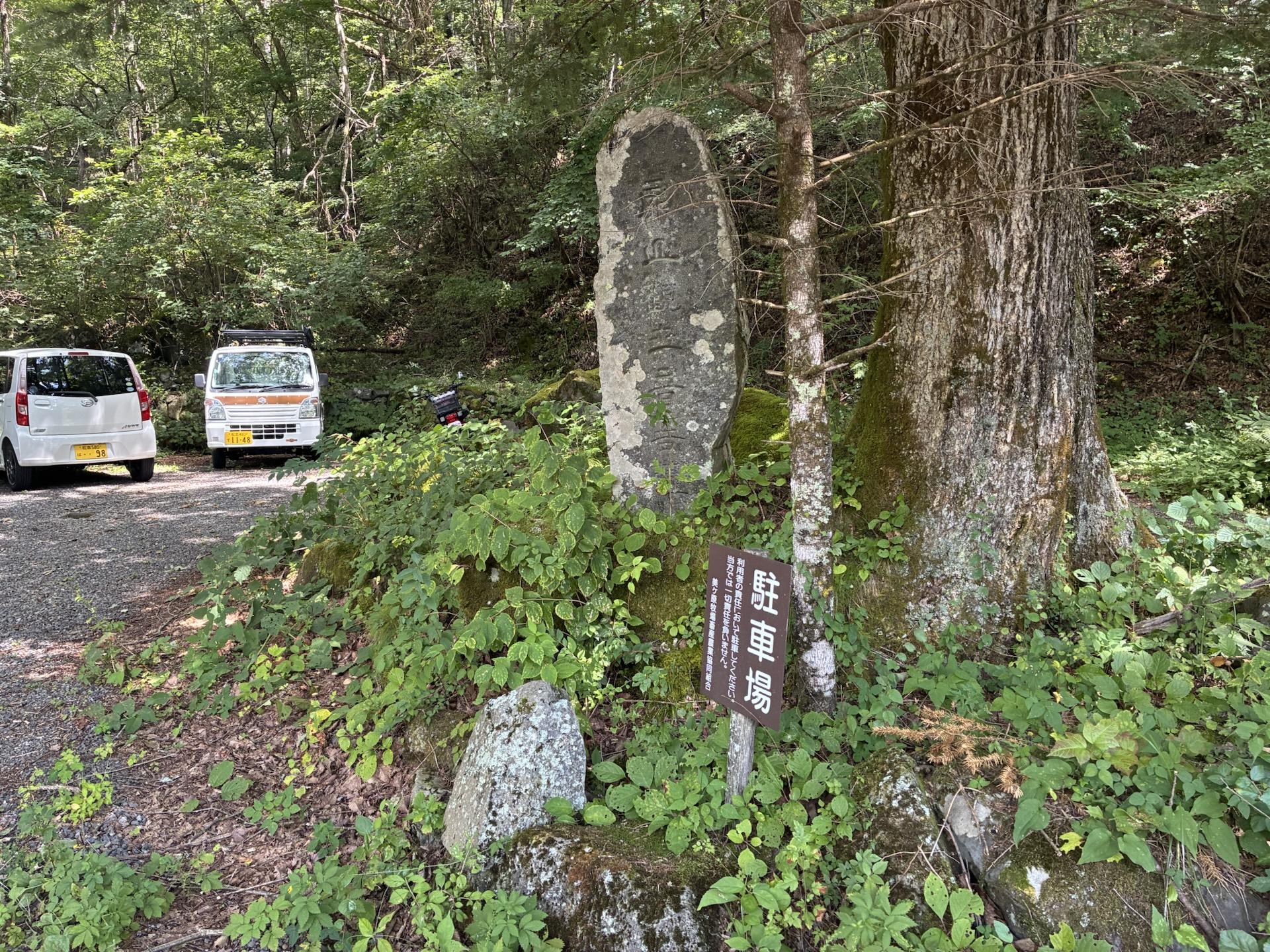 三城いこいの広場の駐車場。奥に数台の軽自動車が停まり、手前には「駐車場」と書かれた木の標柱が立っている。周囲は緑の樹林