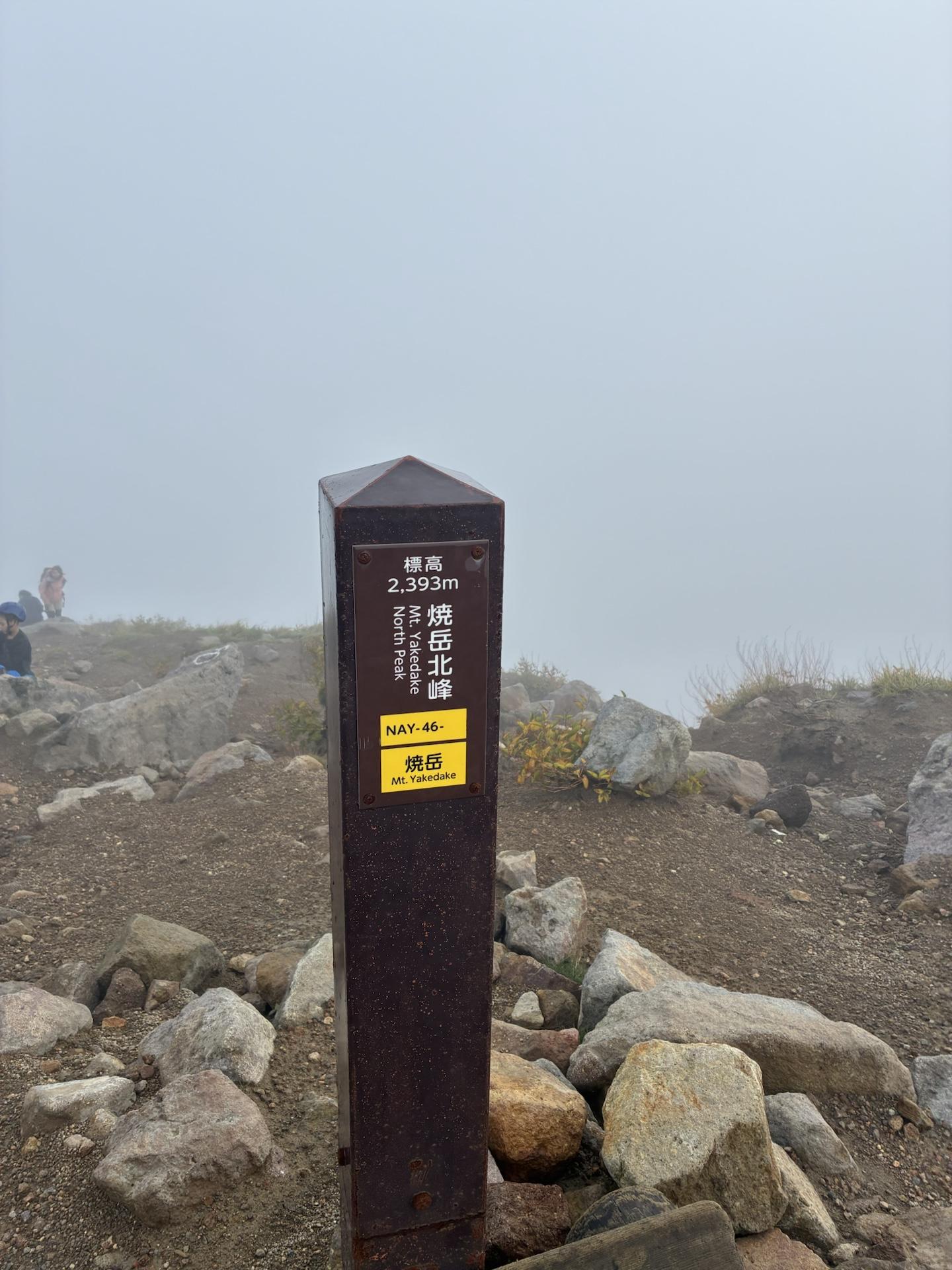 焼岳北峰の山頂標識。標高2,393mと書かれた木製の柱が立ち、背景は一面の濃いガスで何も見えない