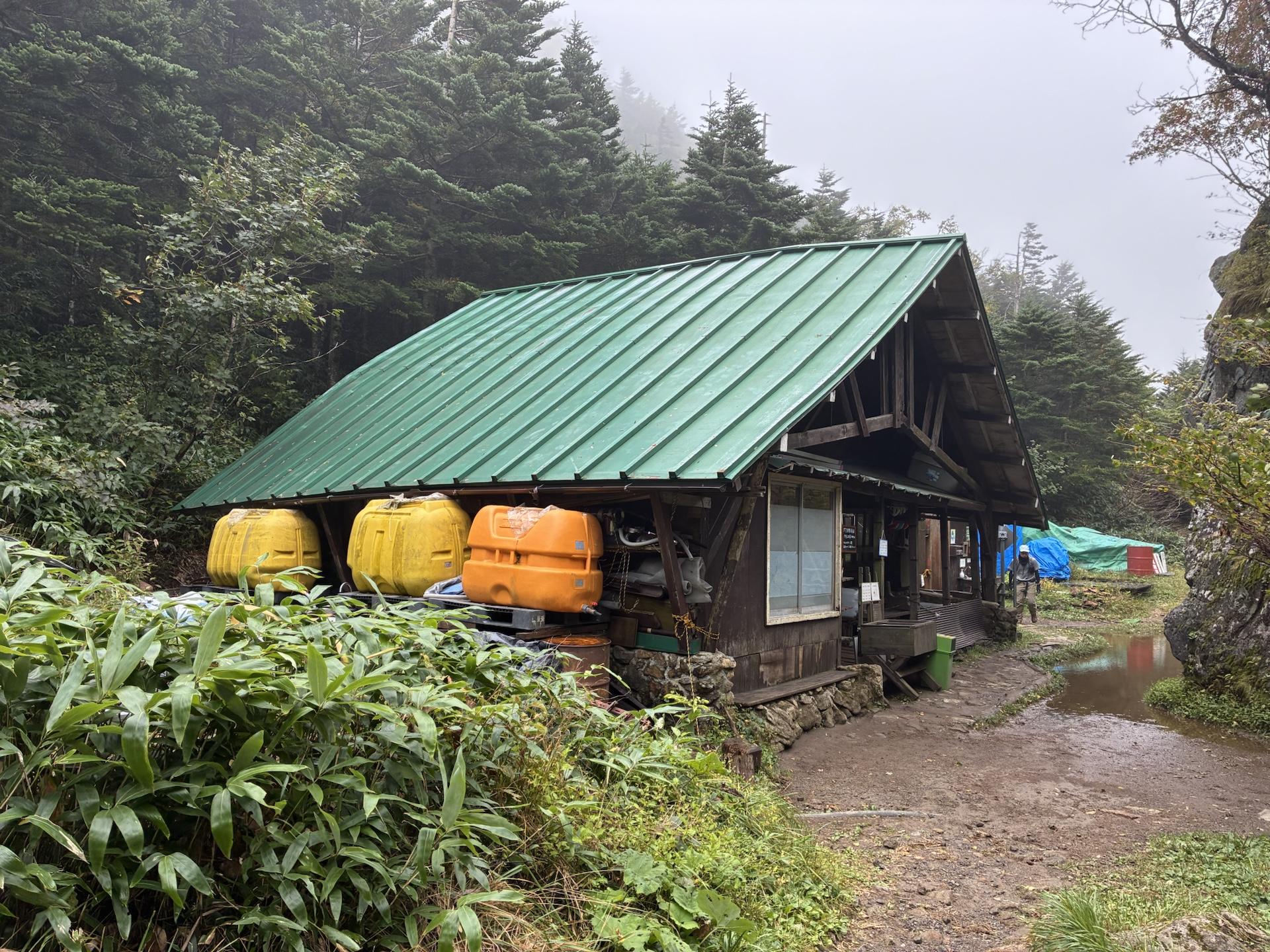 ガスの中に建つ焼岳小屋。緑色の屋根の木造小屋で、軒下に黄色とオレンジの貯水タンクが並んでいる