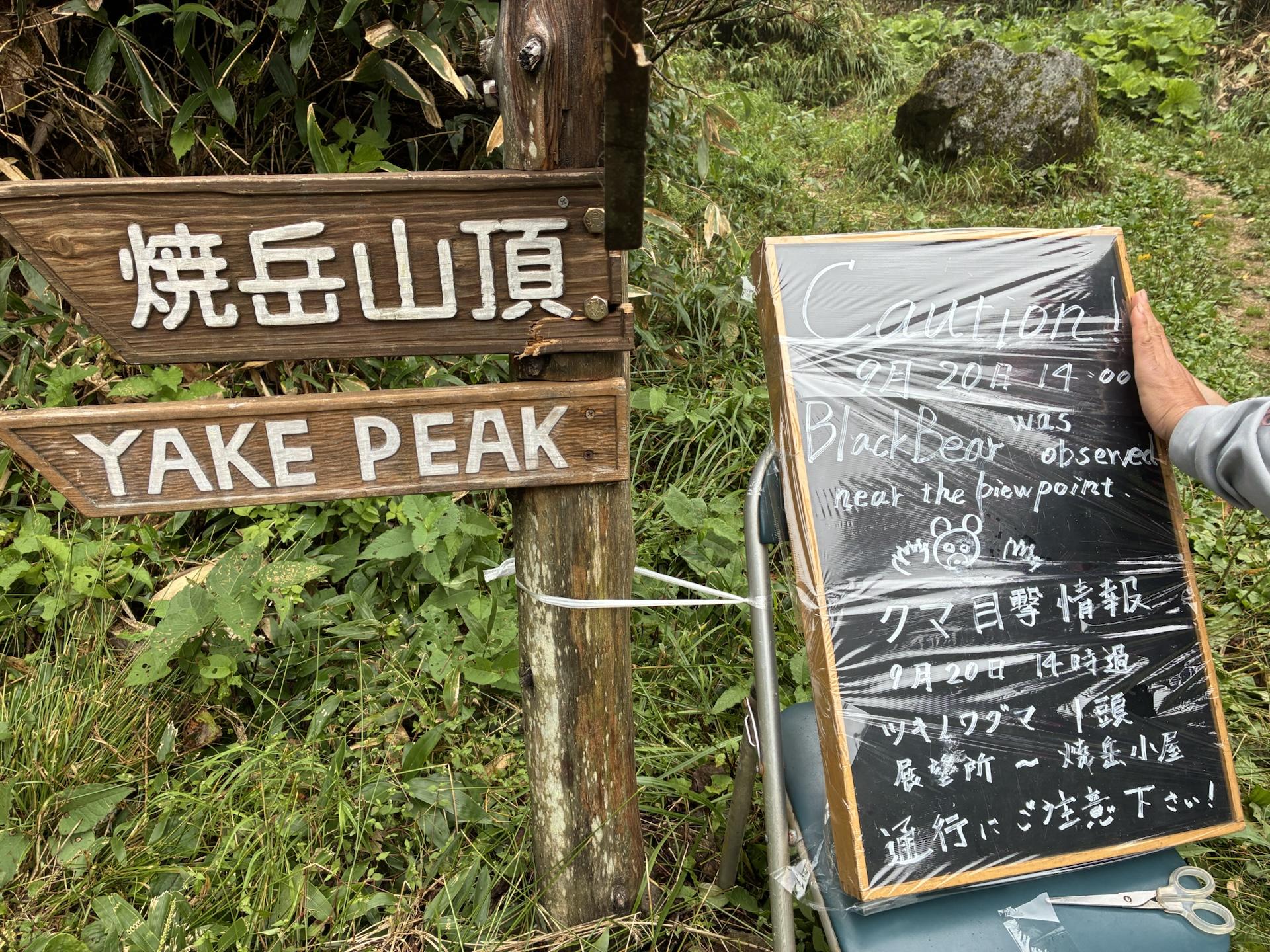 「焼岳山頂 YAKE PEAK」の木製道標の隣に、クマ目撃情報を知らせる黒板が置かれている。9月20日14時頃、展望所から焼岳小屋の間でツキノワグマ1頭を目撃と書かれている