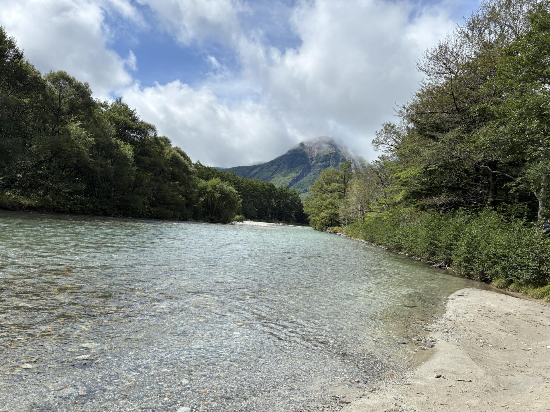梓川の河原から見上げた焼岳。透明な浅瀬の向こうに緑の樹林が続き、その奥に山頂へ雲をかけた焼岳がそびえる