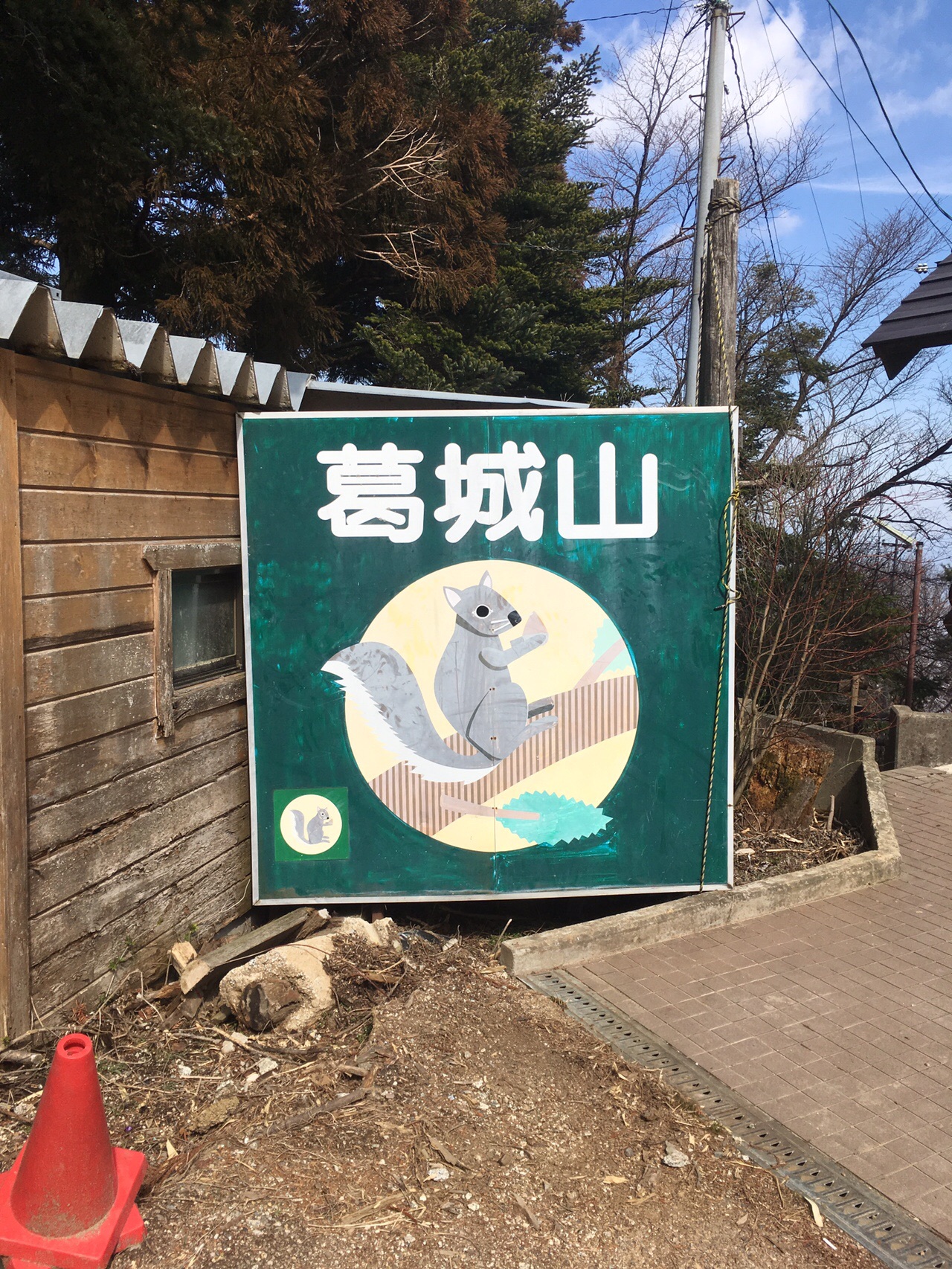 葛城山の緑色の案内看板。リスのイラストが描かれている。奥は木造の建物と青空