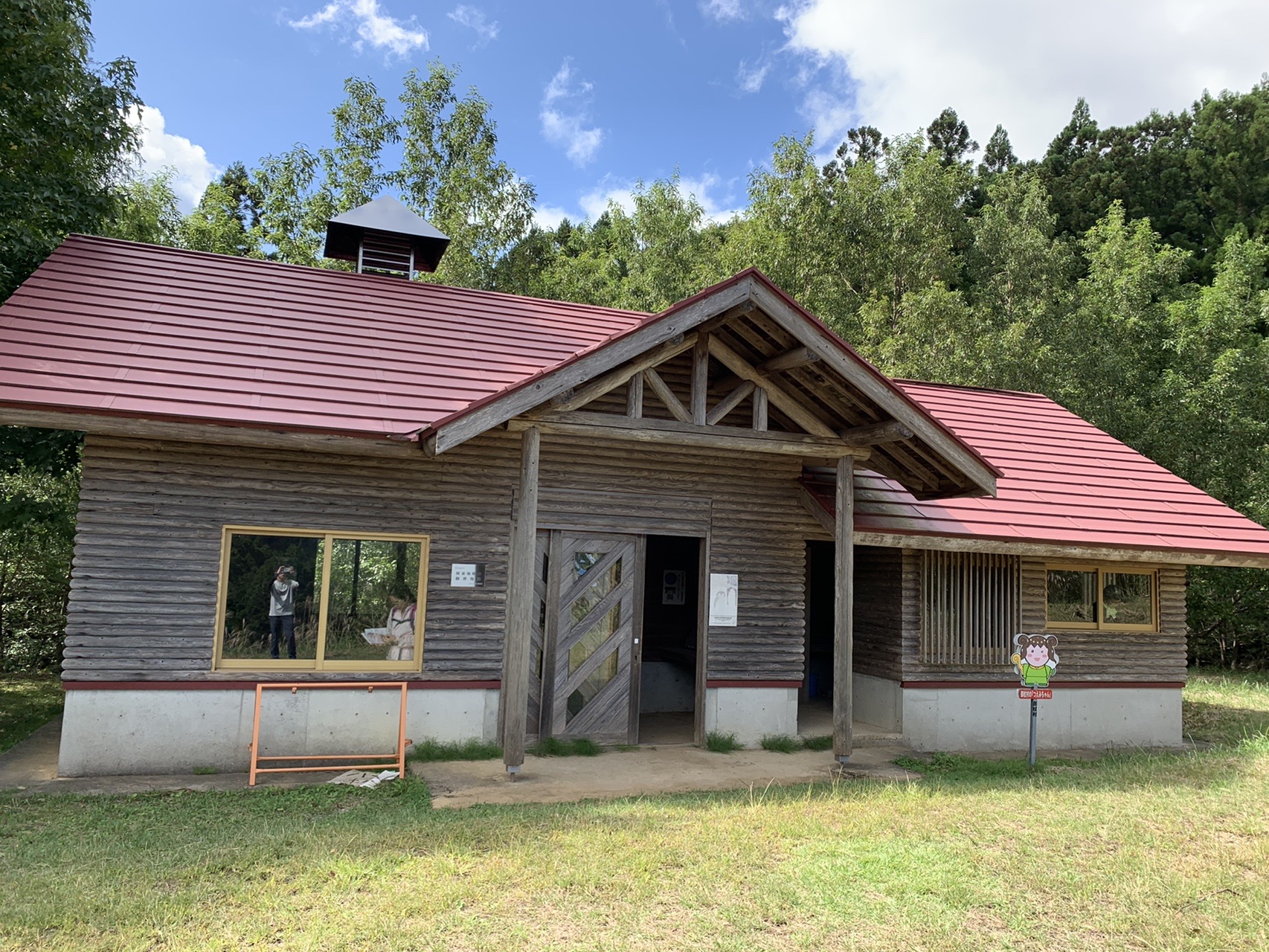 赤い屋根の木造の山小屋。青空と白い雲を背景に草地に建っている