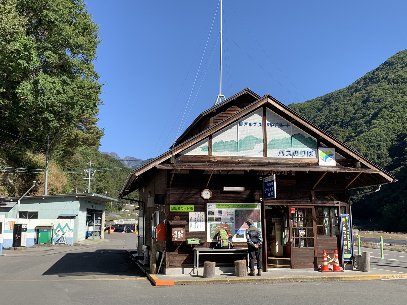 山あいに建つ木造の南アルプス林道バスのりば。青空の下、前に駐車スペースが広がっている