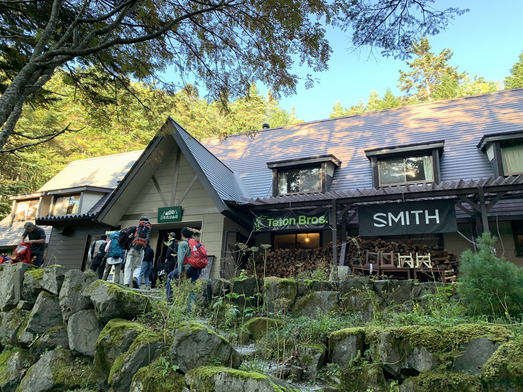 緑と灰色の屋根の山小屋。木立に囲まれ、前に登山者が数人立っている