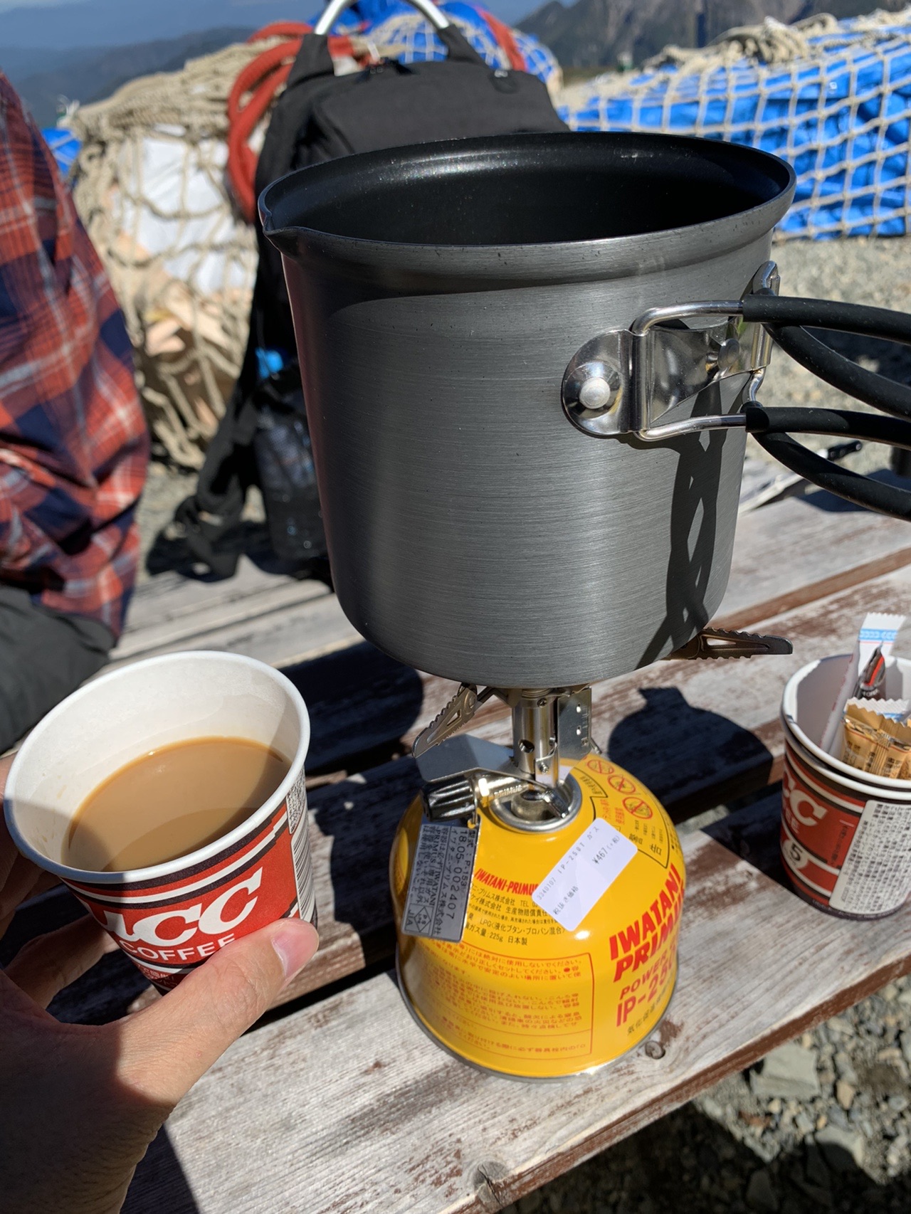 山小屋のベンチに置かれたバーナーとクッカー。手前に紙コップに入ったコーヒーがある
