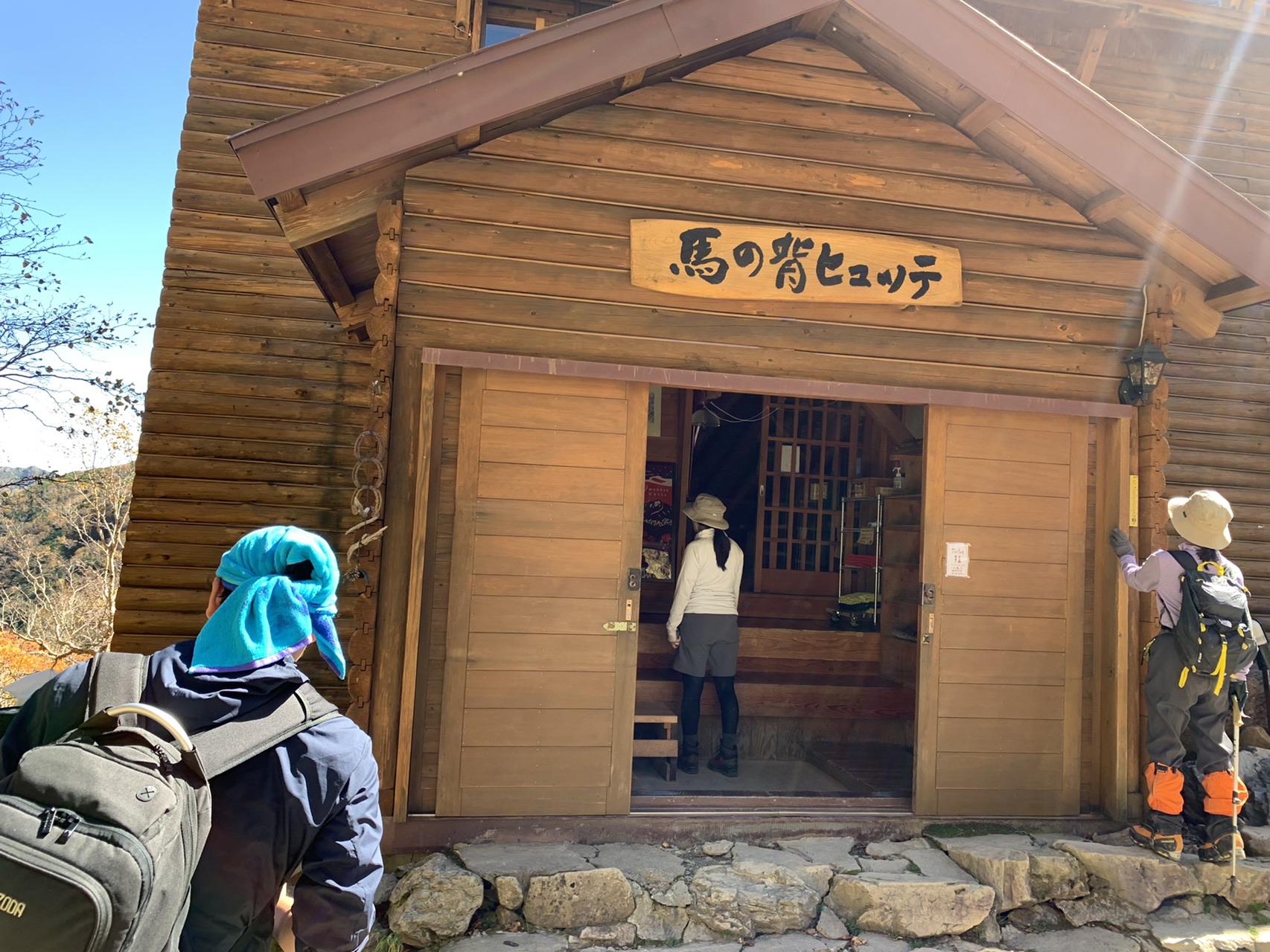 木造の山小屋の入口。「馬ノ背ヒュッテ」と書かれた看板が掲げられ、登山者が出入りしている