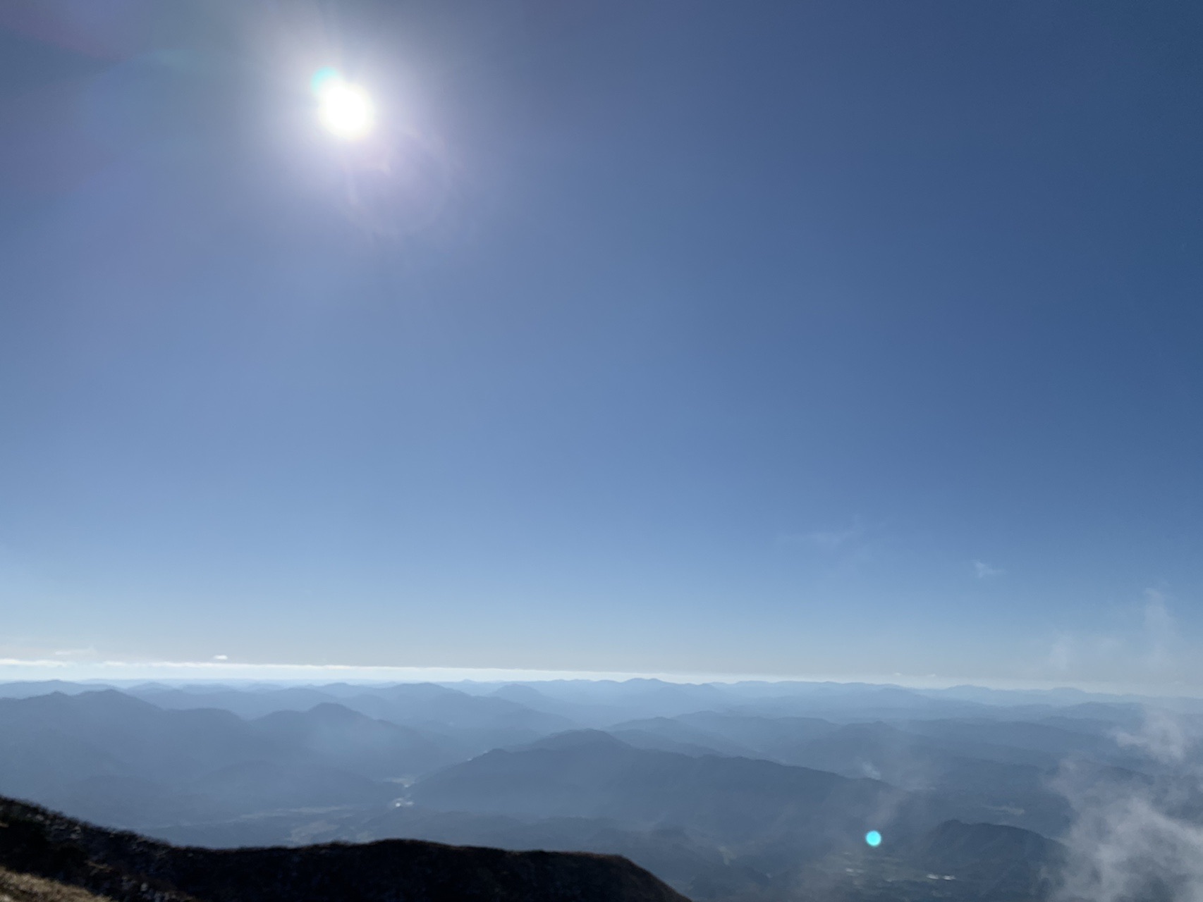 太陽が高く輝き、その下に雲海が水平に広がっている。手前は山の斜面のシルエット
