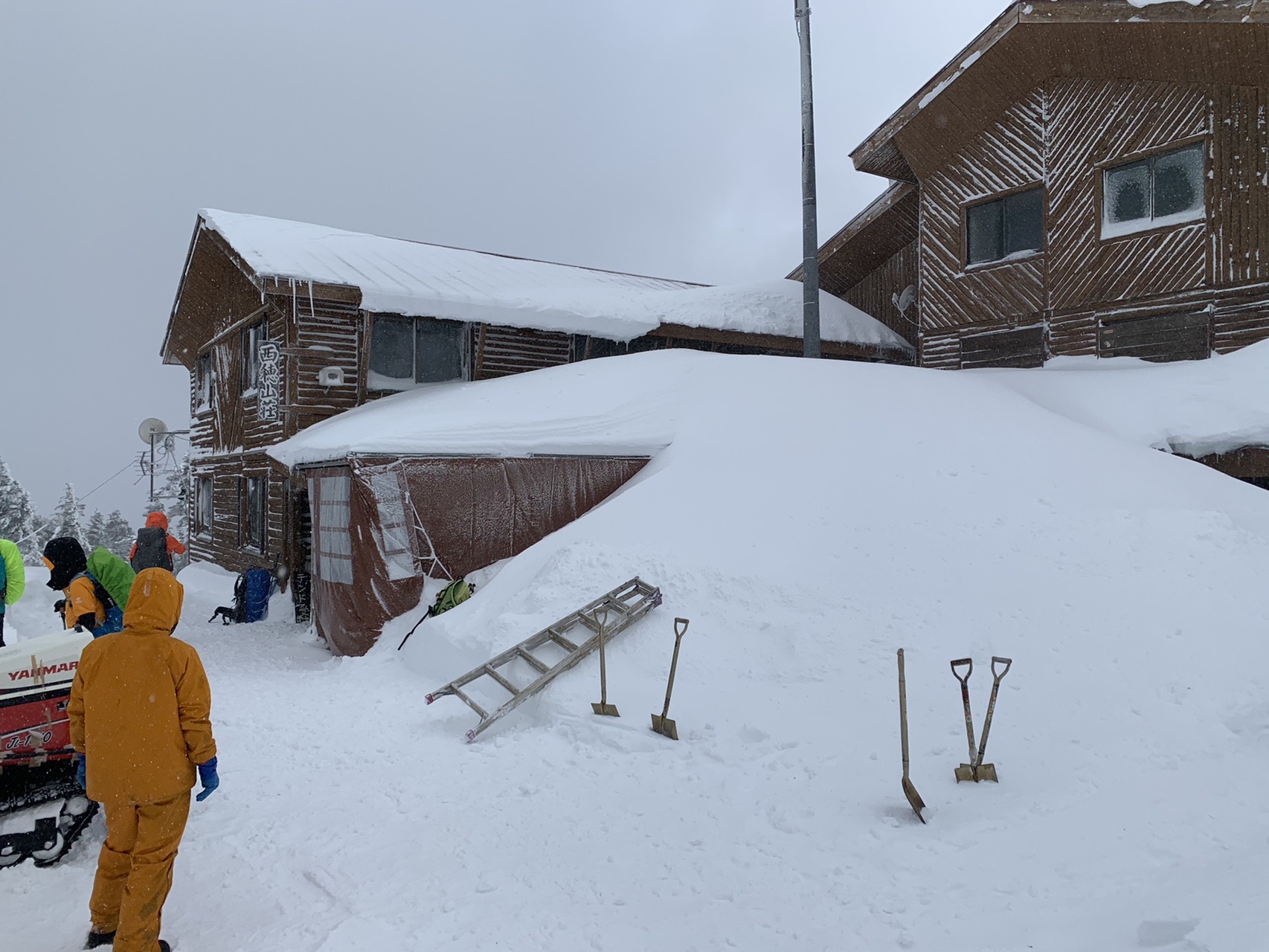 屋根まで雪に覆われた木造の山小屋。前の雪面にスコップが刺さっている