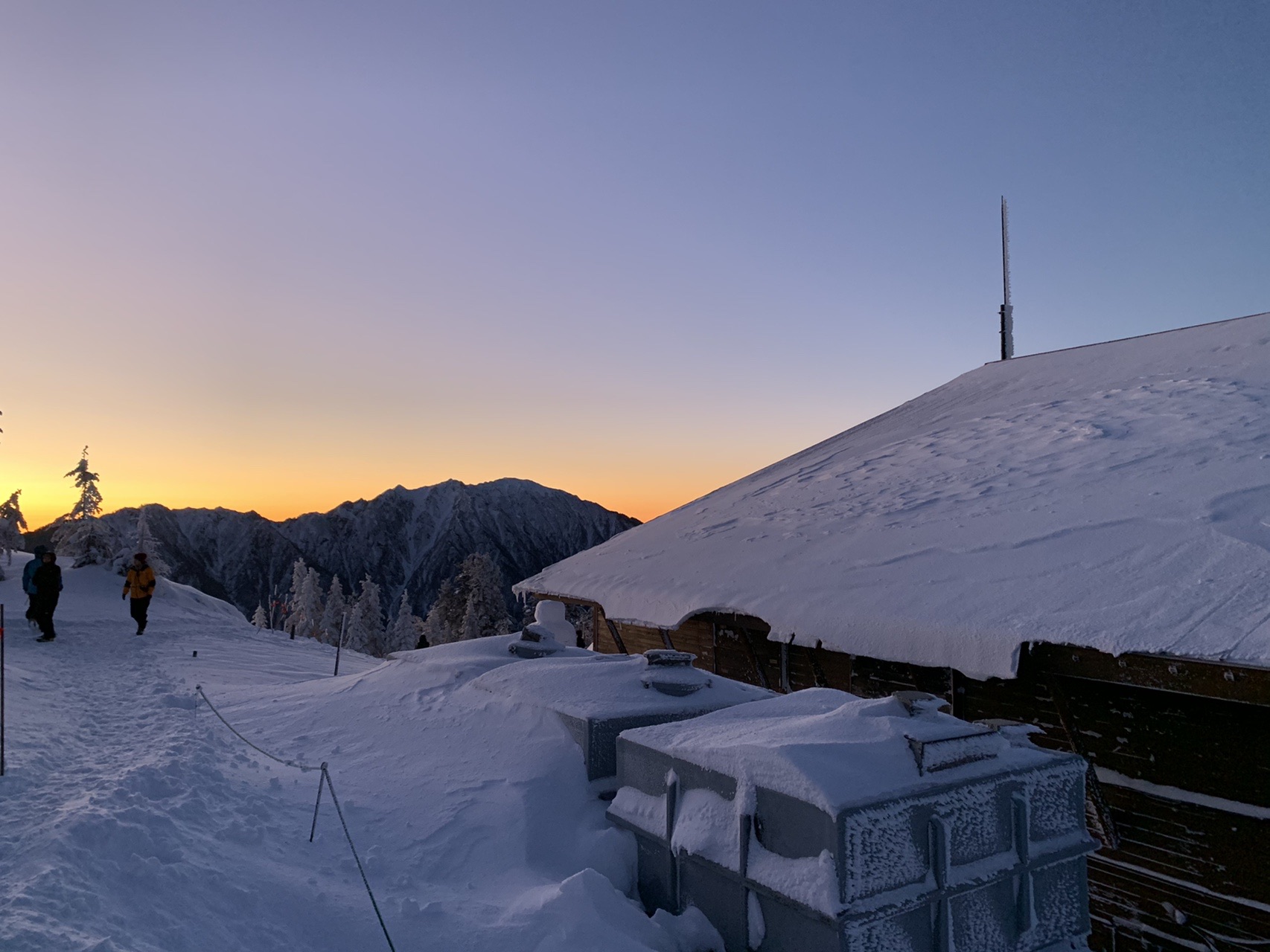 雪に埋もれた山小屋の屋根越しに、オレンジ色に染まった空と山の稜線が見えている。左に電波塔のようなポールが立つ