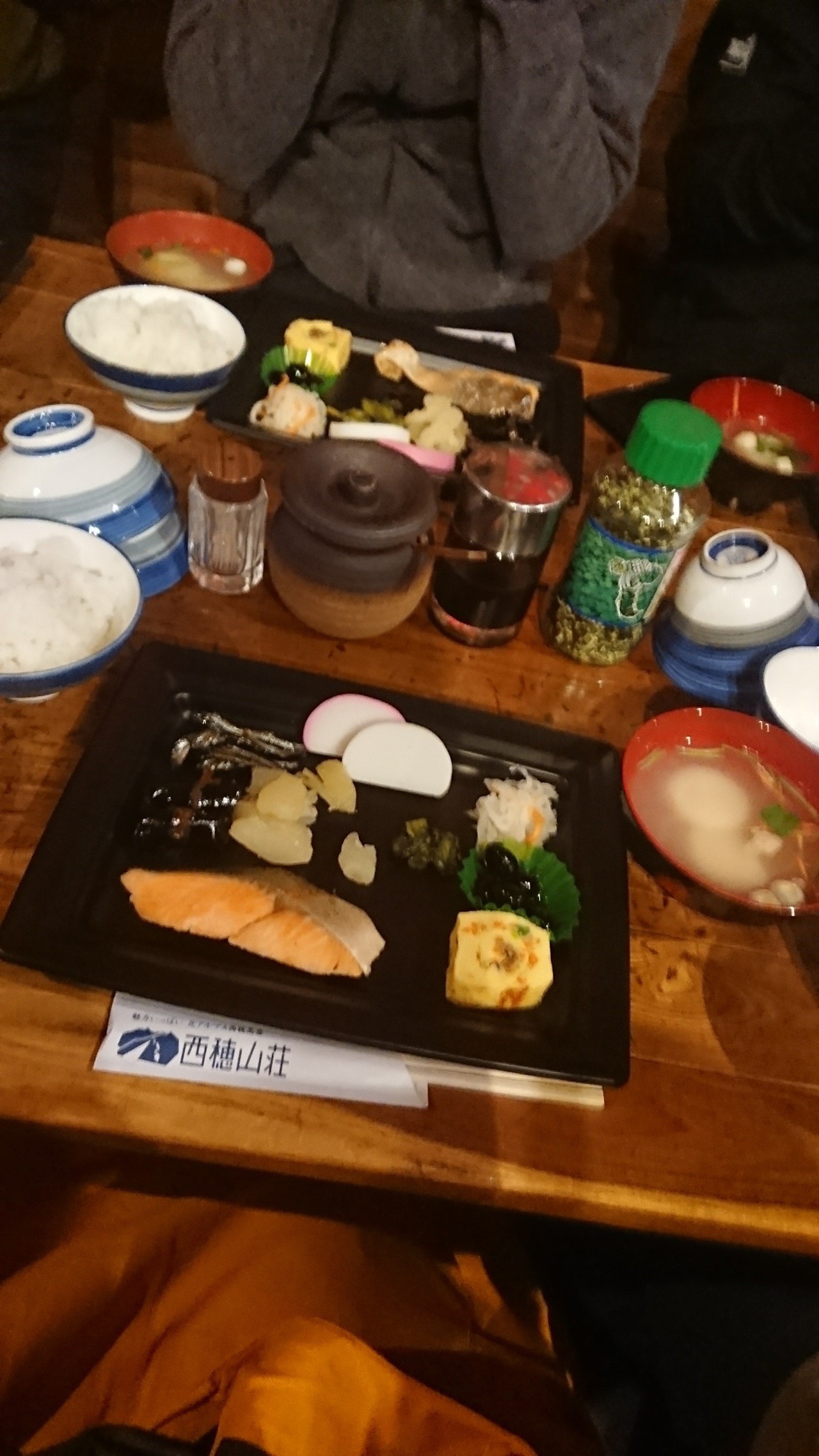 山小屋のテーブルに並んだ朝食。黒い重箱におせち料理が詰められ、焼き鮭、ごはん、味噌汁、小鉢が添えられている