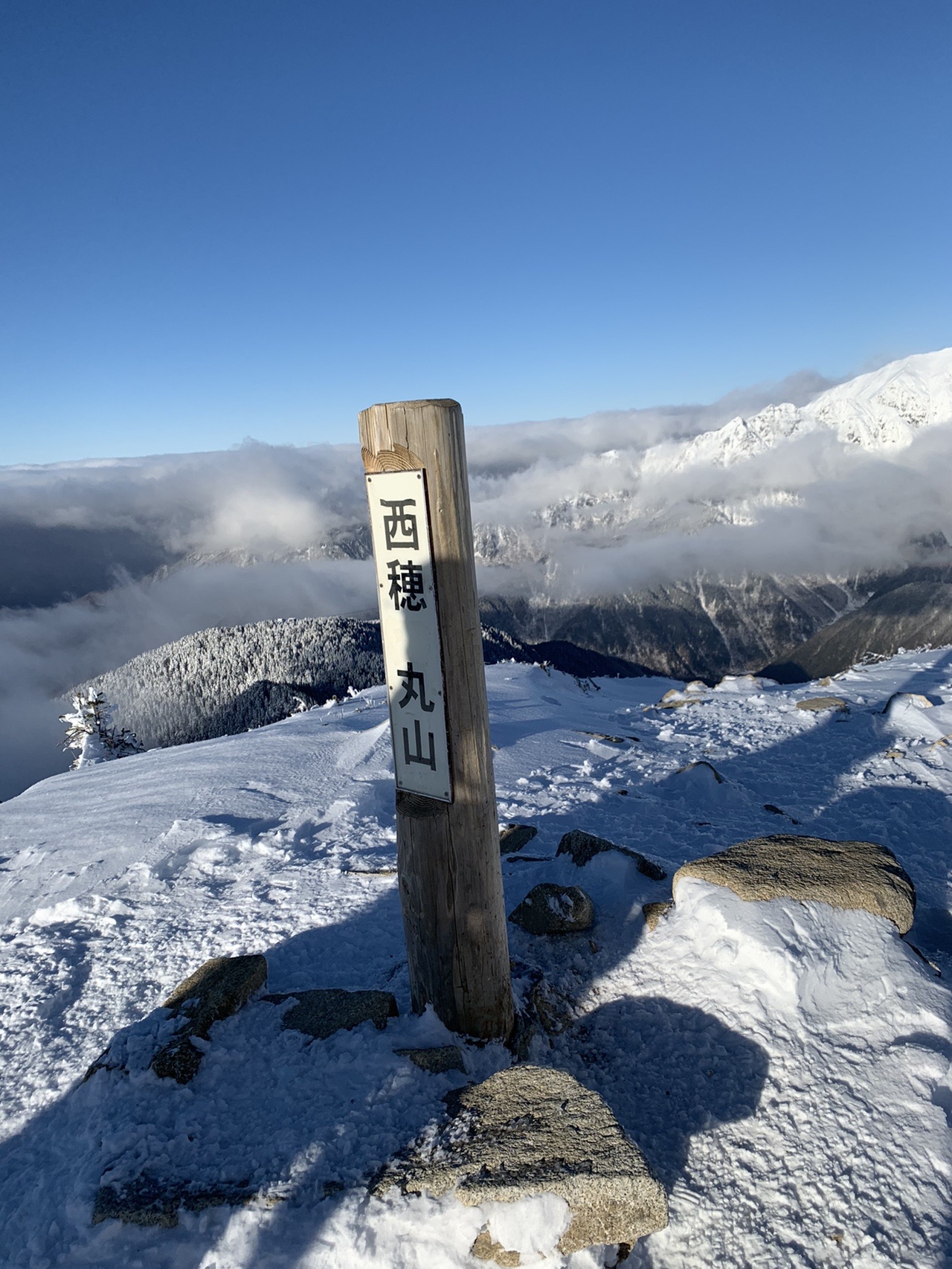 雪原に立つ木の標柱。「丸山」と縦書きされている。奥に雪の稜線と雲海が広がる