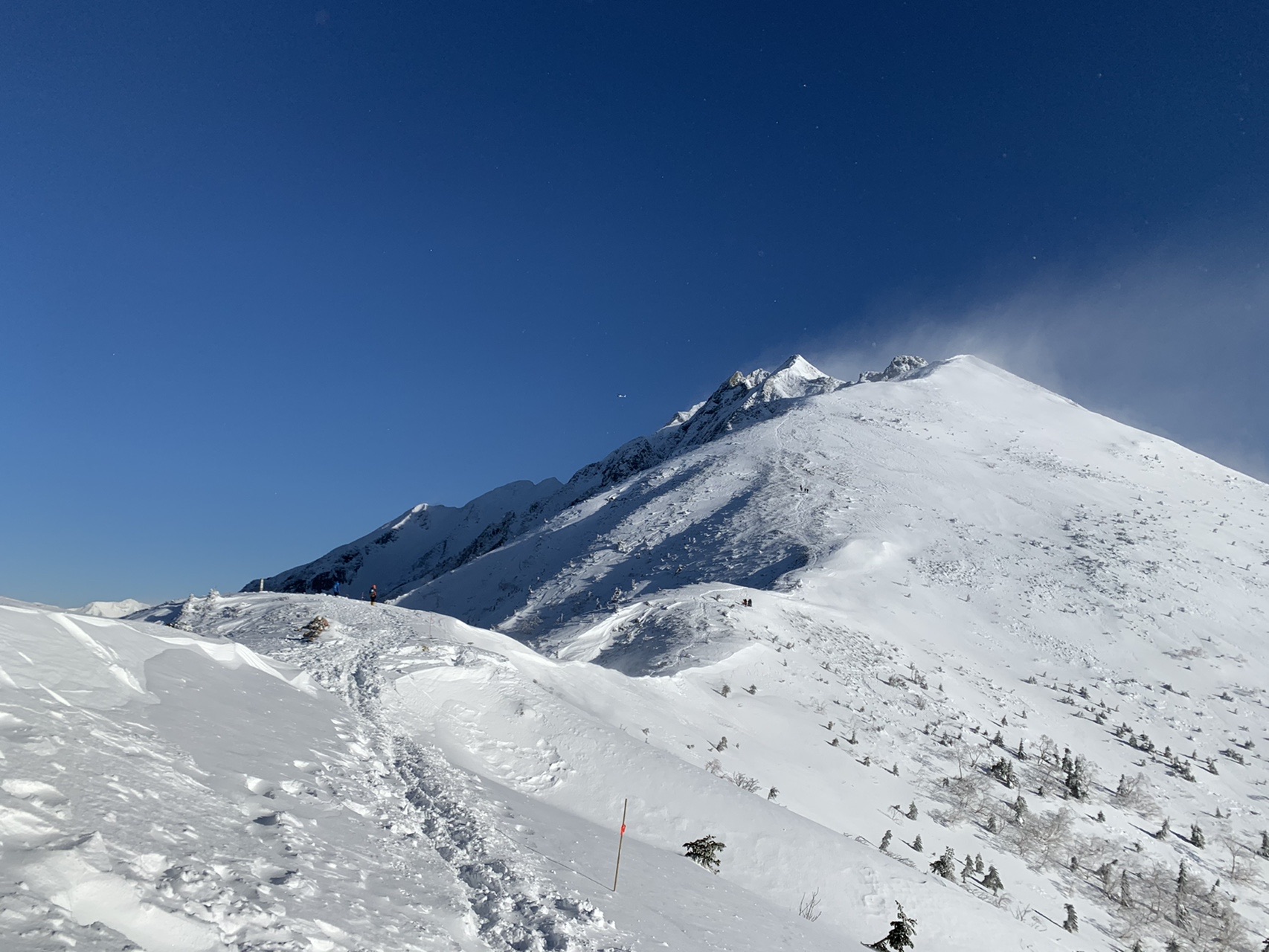 真っ青な空の下に伸びる雪の稜線。手前の雪面に足跡が続き、奥にとがったピークが連なる