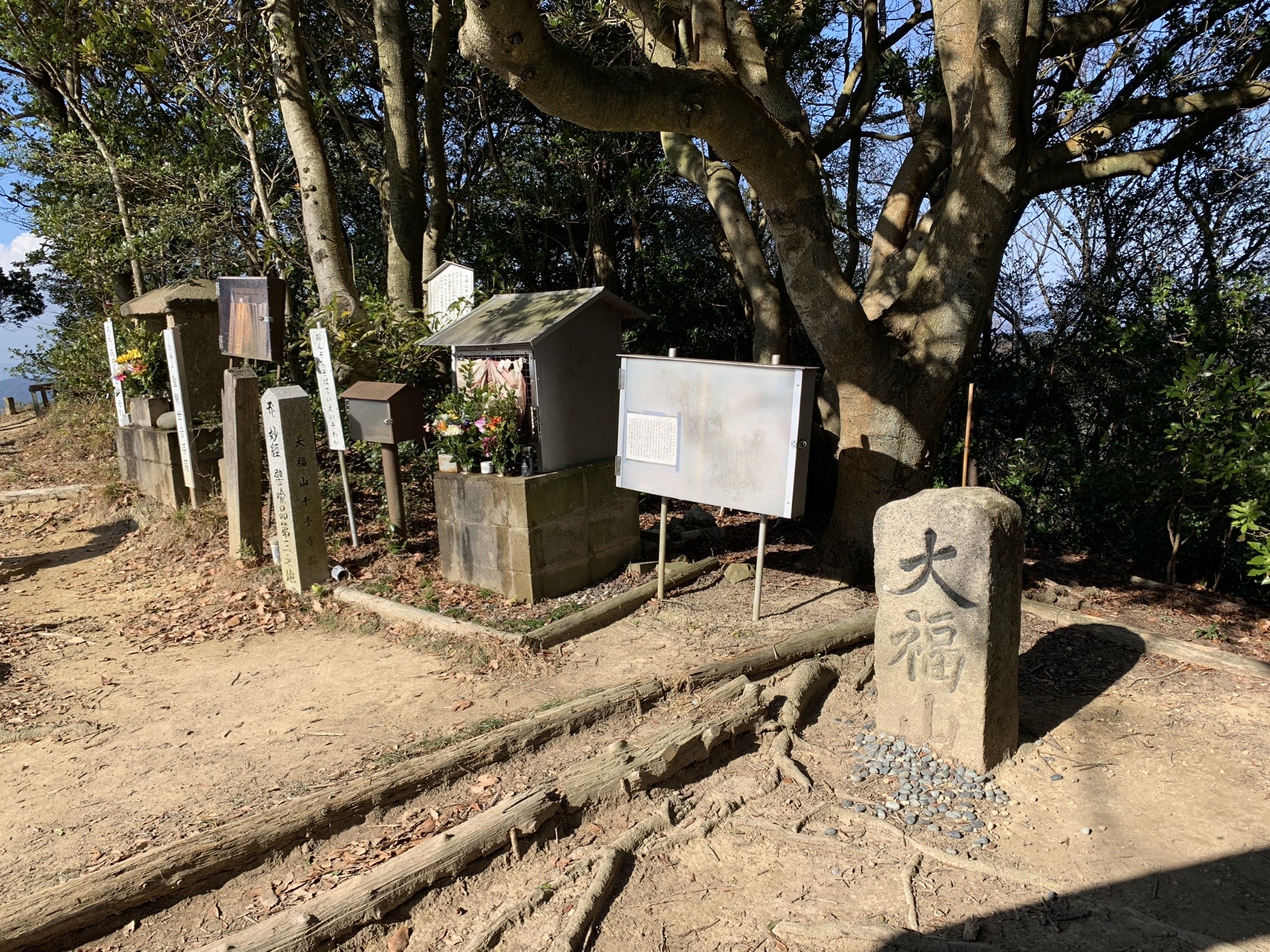 山中に立つ古い石柱と、その横の小さな祠、白い説明板。石柱には「大福山」と刻まれている
