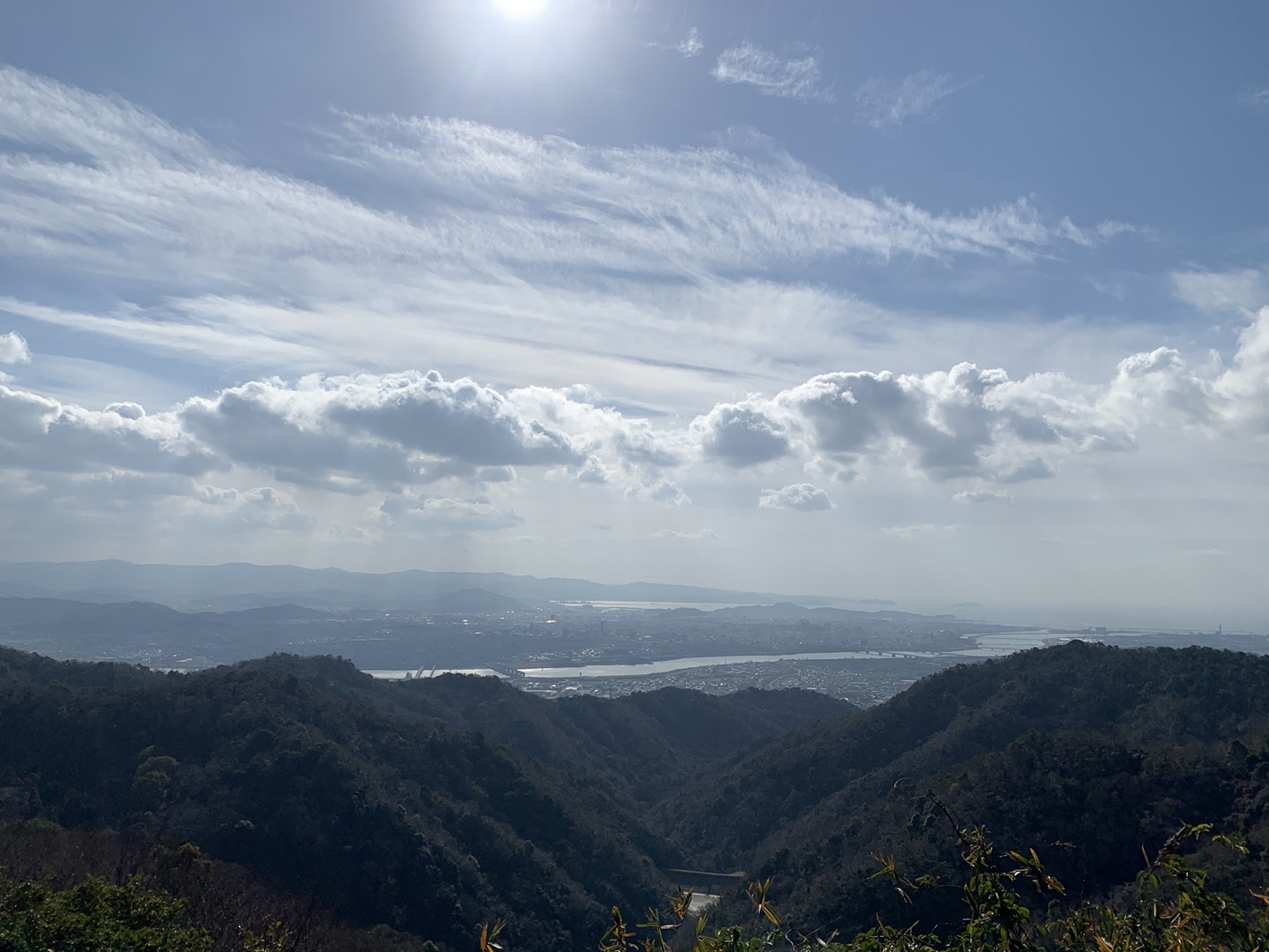 尾根から見下ろす和歌山側。太陽が高く、雲が低く広がり、その下に川と市街地が見えている