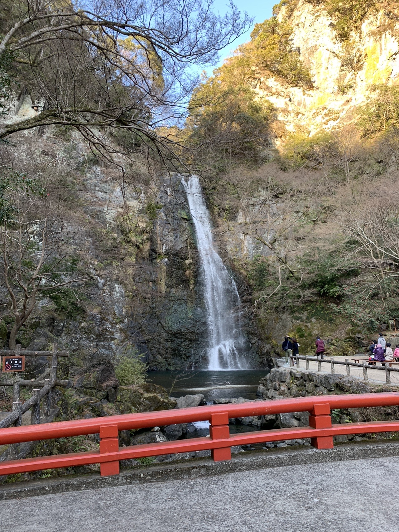 岩壁を落ちる大きな滝。手前に赤い欄干の橋が架かり、両側を木々に囲まれている