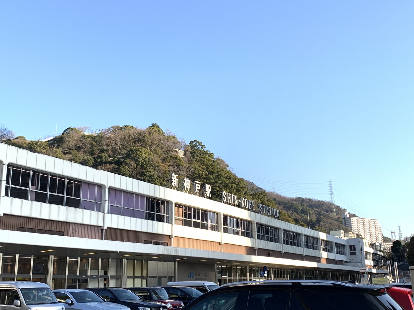 新神戸駅の駅舎を下から見上げた写真。高架の駅の下に駐車場が並び、青空が広がっている
