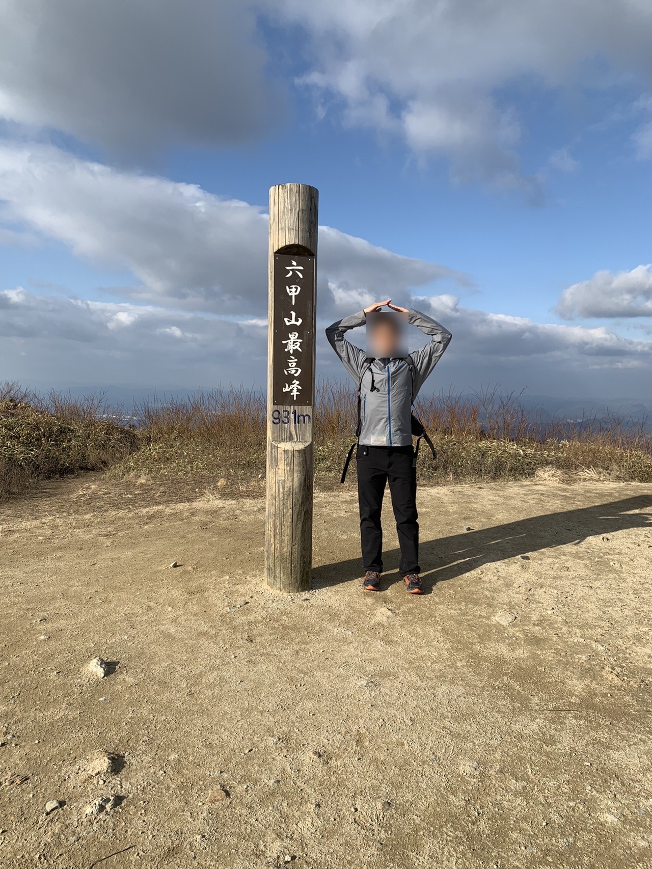 「六甲山最高峰 931m」と彫られた木の標柱の横で、両腕を頭の上で三角にして山のポーズをとる人