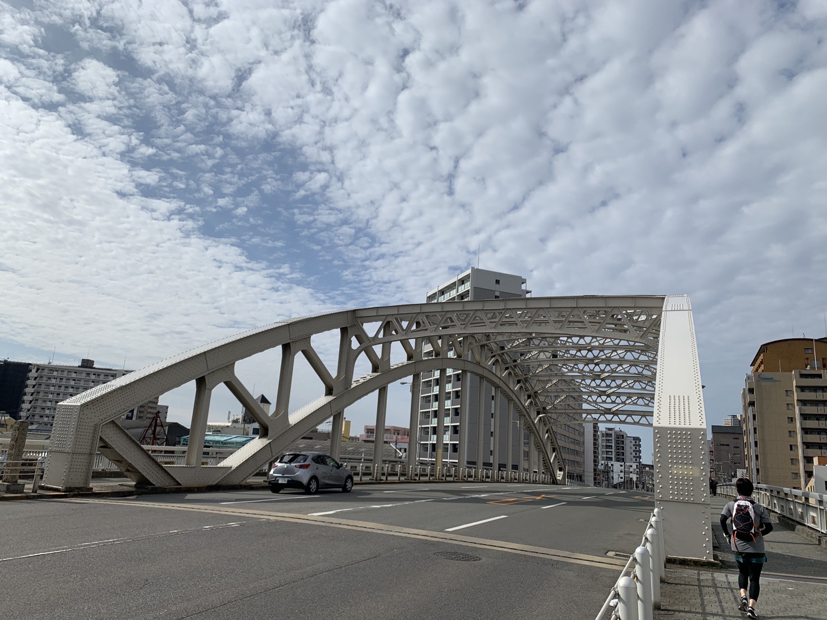 道路をまたぐ鉄骨のアーチ橋。歩道が付いていて、車が走っている。空にはうろこ雲