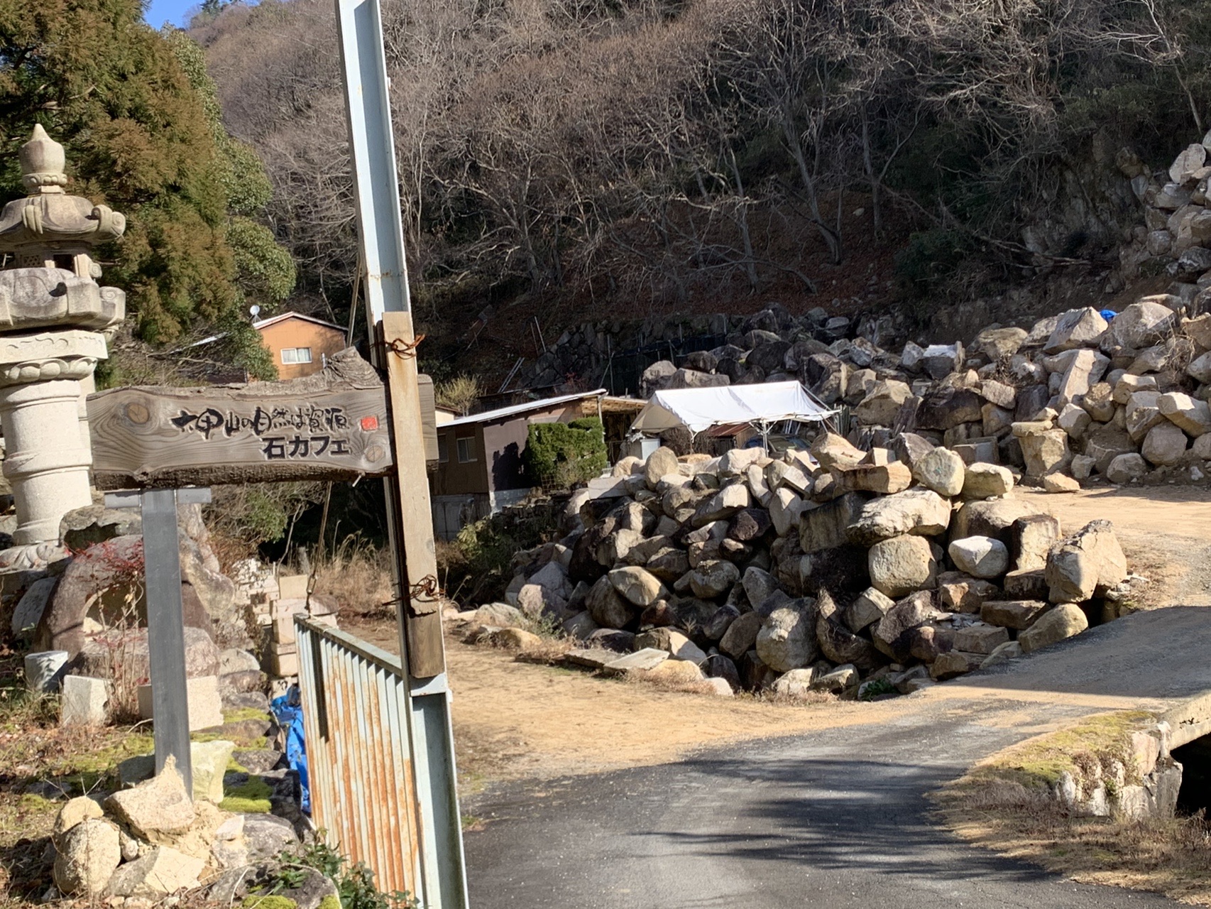 石灯籠のわきに立つ木の看板。「六甲山の自然は絶頂 石カフェ」と手書きで書かれている。奥に積み上げられた石材
