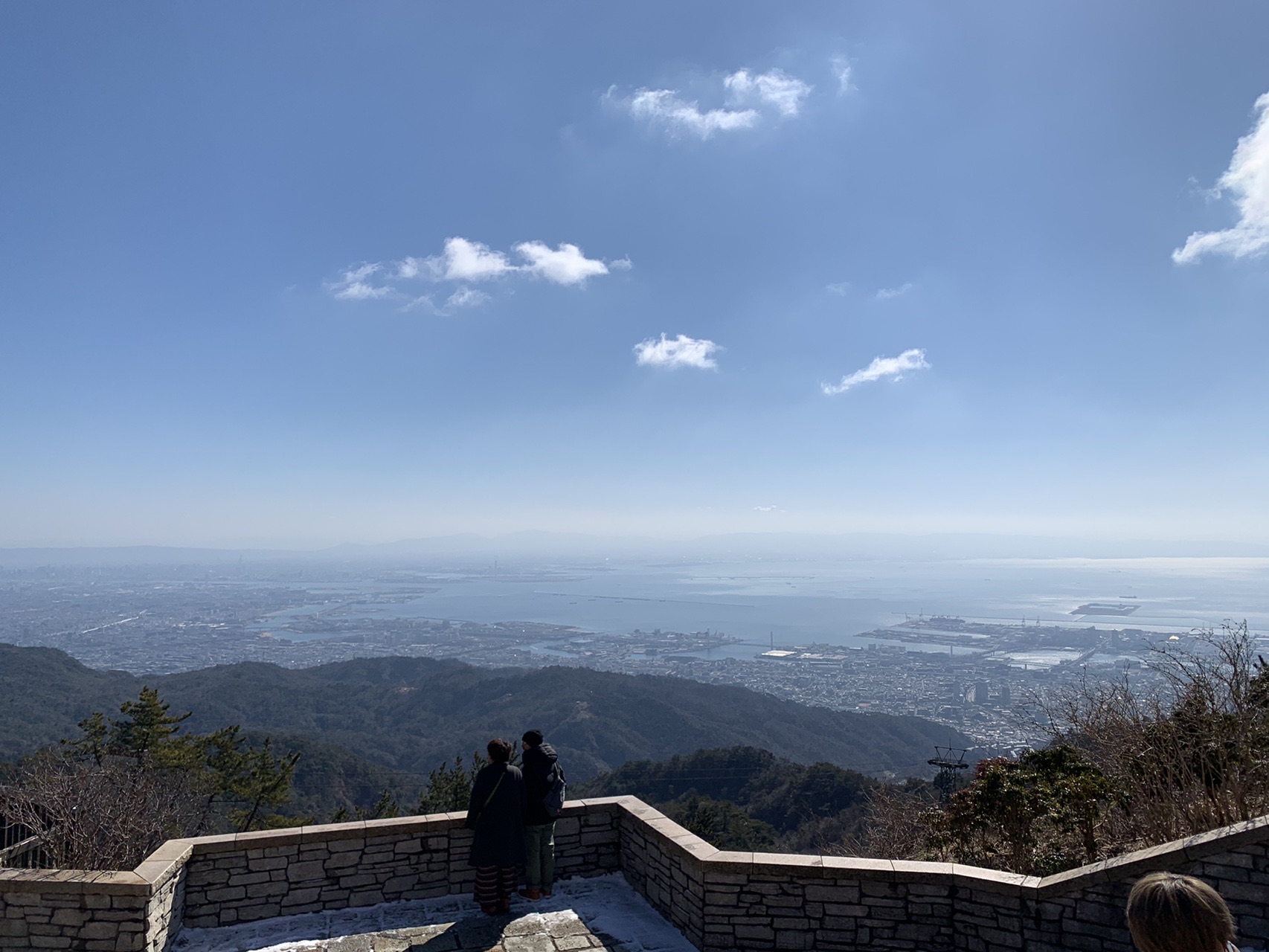 石積みの展望台から見下ろす神戸の街と大阪湾。かすんだ海に沿って市街地が広がり、手前に2人が立っている