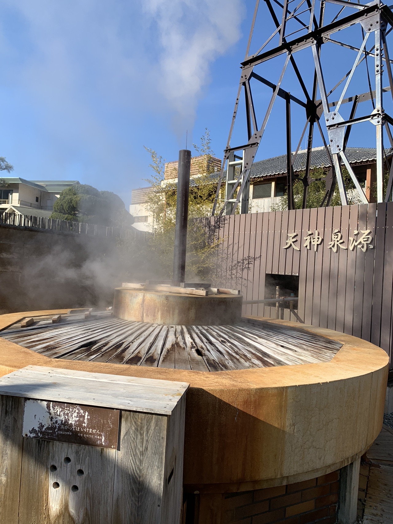 温泉街の源泉。丸い木製の蓋から白い湯気が勢いよく立ち上り、後ろに「天神泉源」と書かれた壁と櫓が見える