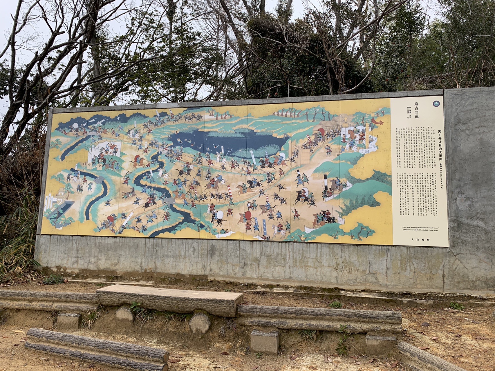 屋外に設置された横長の陶板絵図。合戦の場面が描かれ、右側に解説文が添えられている