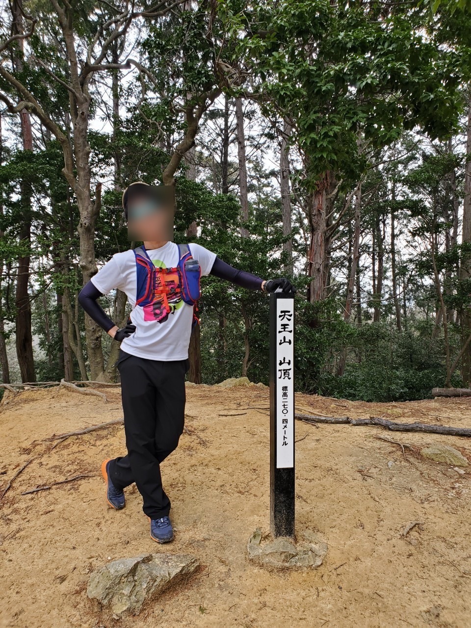 杉林に囲まれた広場に立つ黒い角柱の標識。「天王山 山頂 標高二七〇・四メートル」と縦書きされ、その横にトレイルランナーが立っている