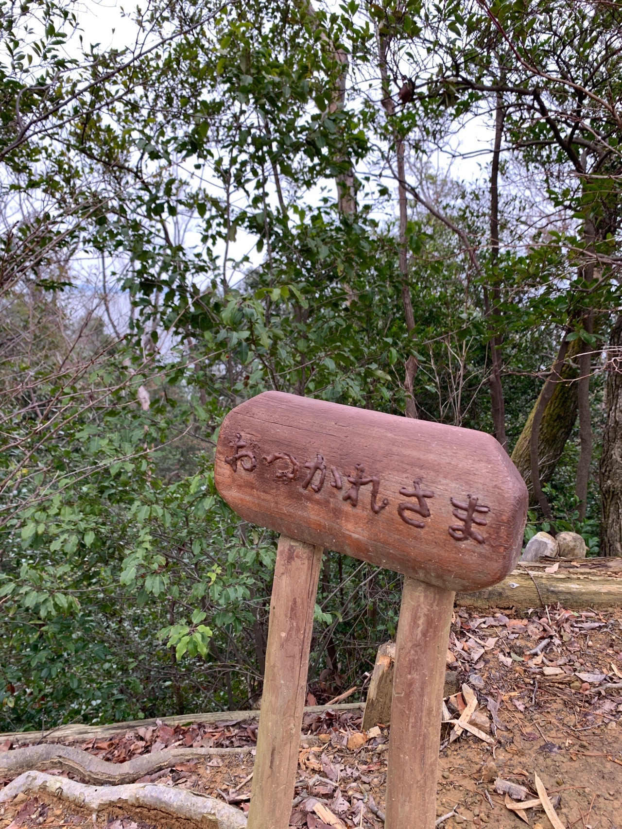 山道のわきに立つ木製の丸い標識。「おつかれさま」と彫り込まれている