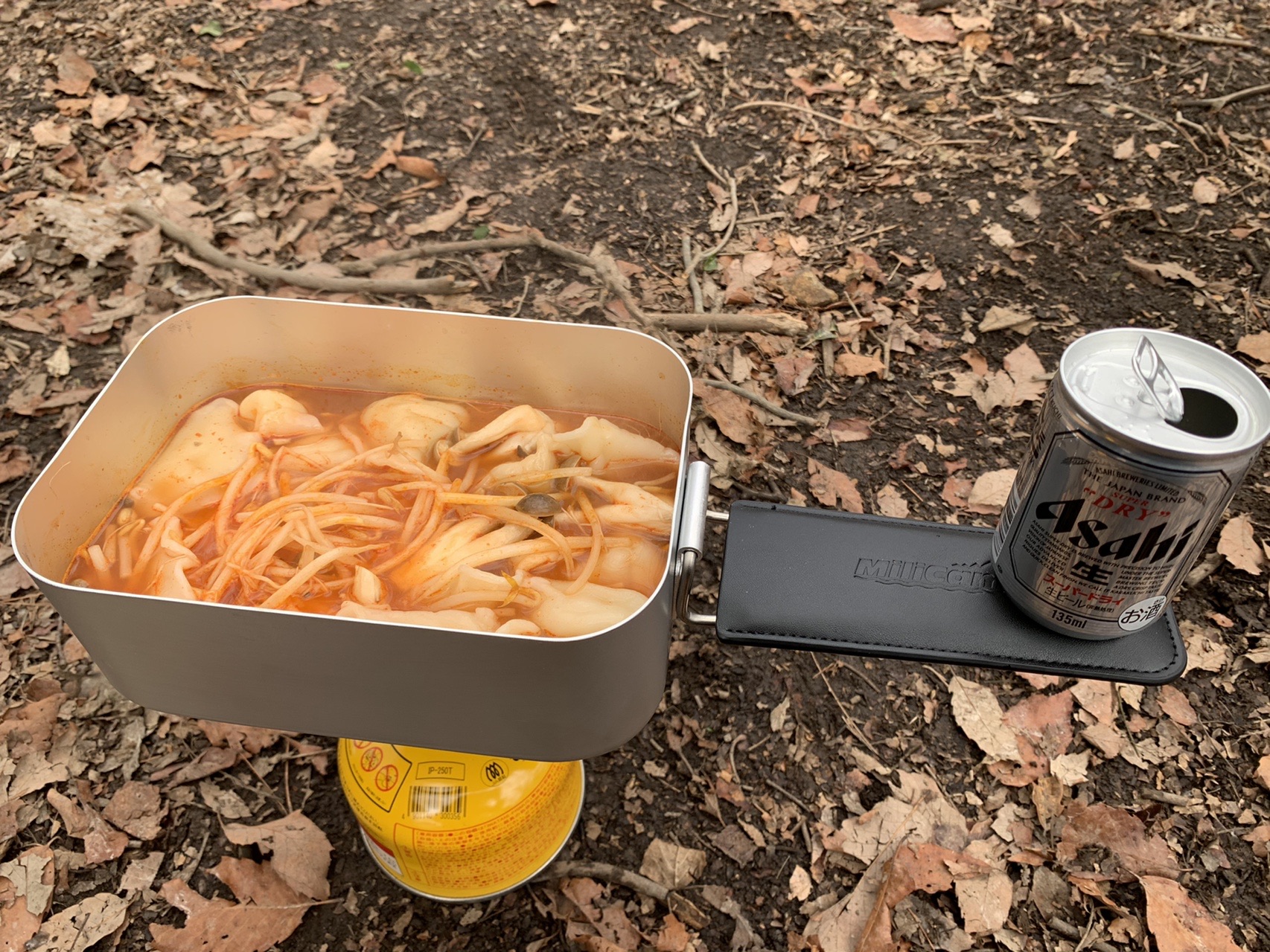 落ち葉の上に置かれた大きな四角いメスティン。中に赤いスープと餃子が入って煮えている。横に缶ビール