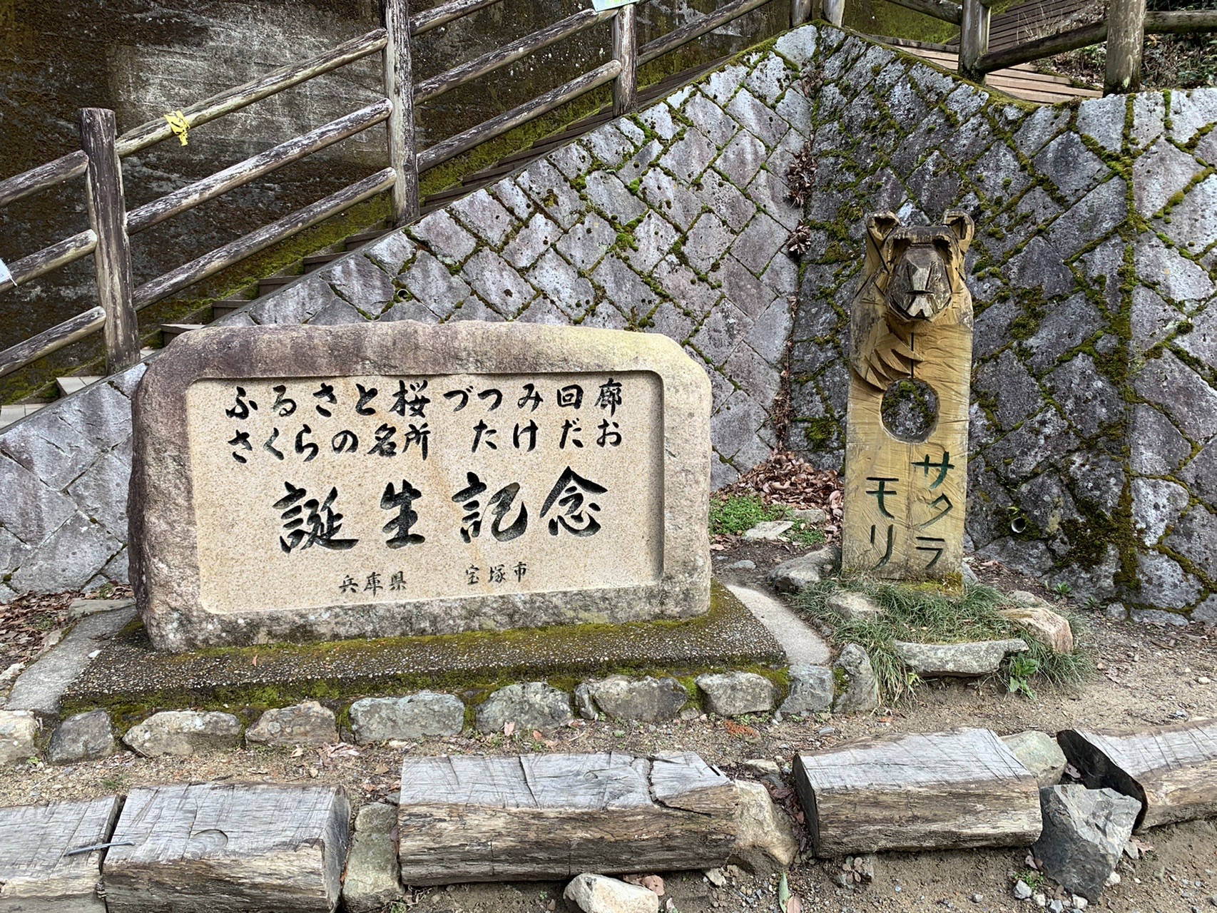石垣の前に置かれた石碑。「ふるさと桜づつみ回廊 さくらの名所 たけだお 誕生記念 兵庫県 宝塚市」と刻まれている。横に木彫りの像が立つ