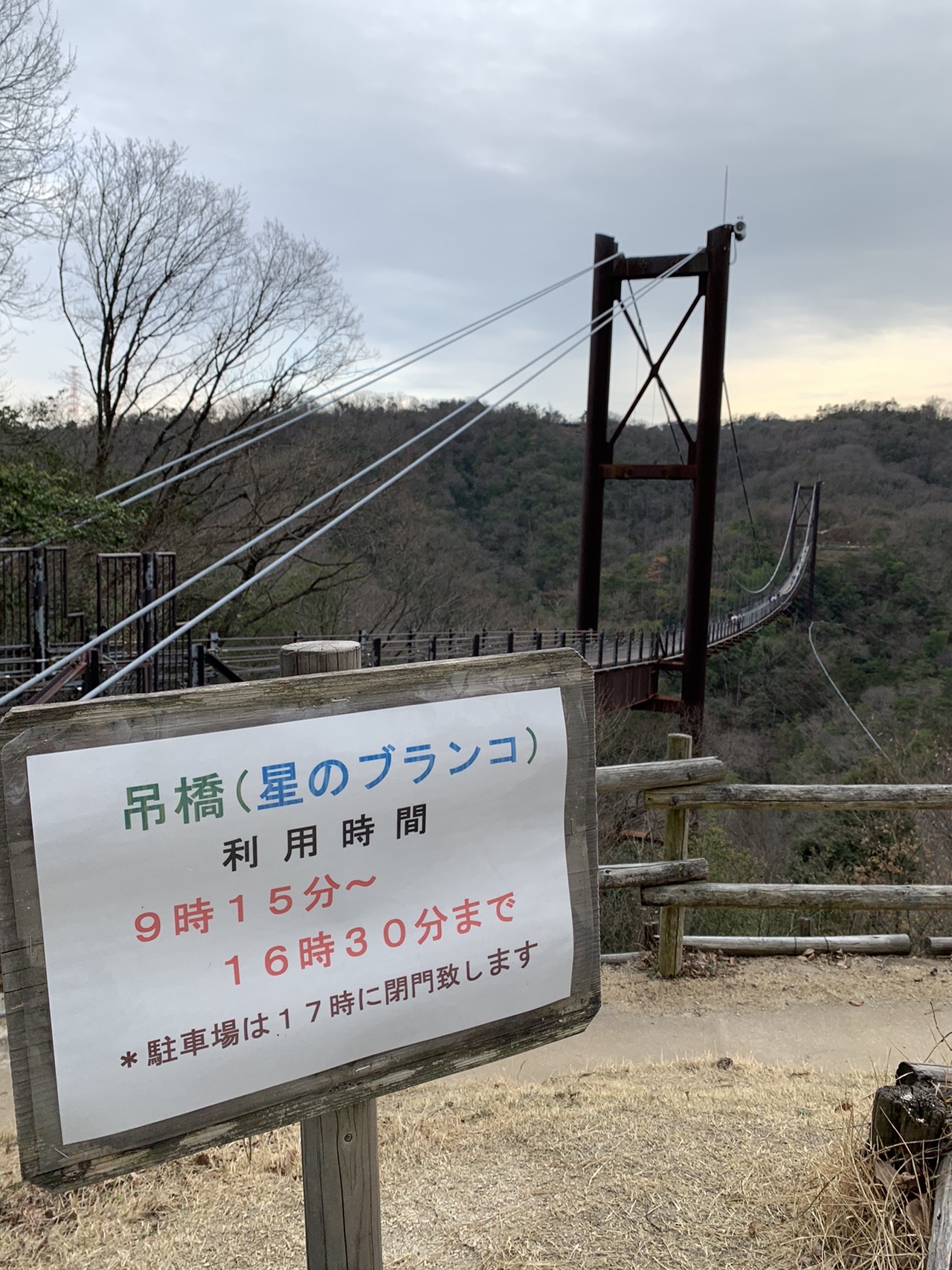 「吊橋(星のブランコ)利用時間 9時15分〜16時30分まで *駐車場は17時に閉門致します」と書かれた看板。奥に吊り橋