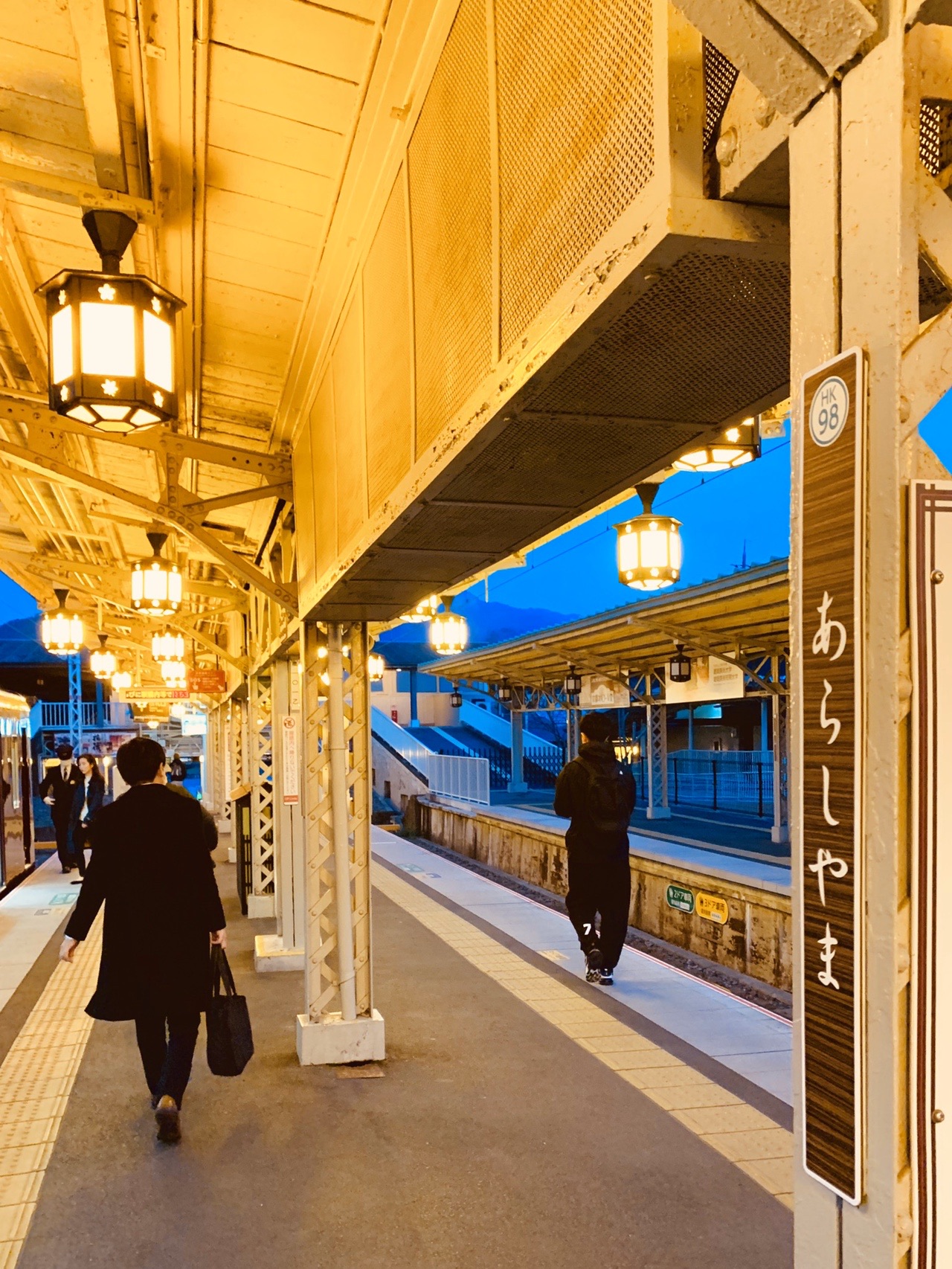 夜明け前の阪急嵐山駅のホーム。木の柱と行灯風の照明が並び、空はまだ青い