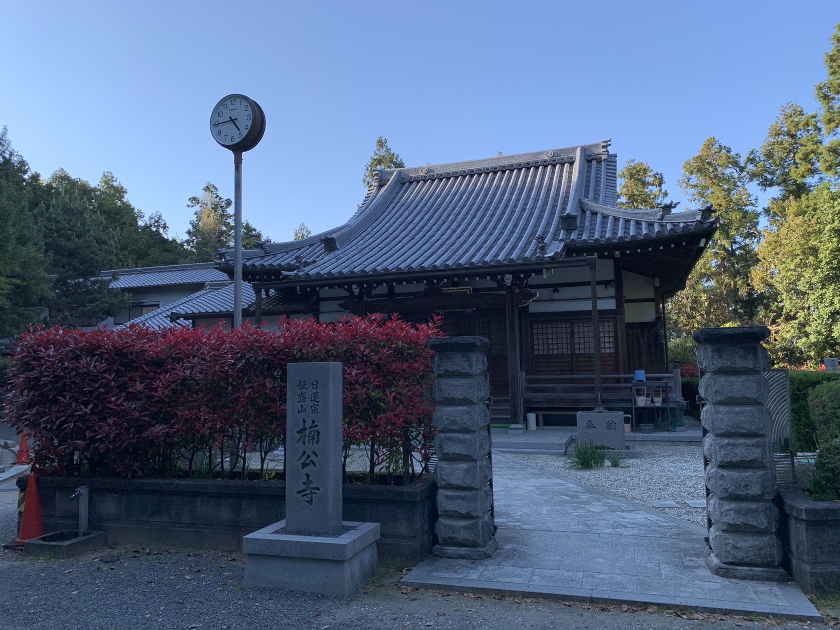 山中の寺。「日蓮宗 飯盛山 楠公寺」と刻まれた石柱と、赤い葉の生け垣、本堂
