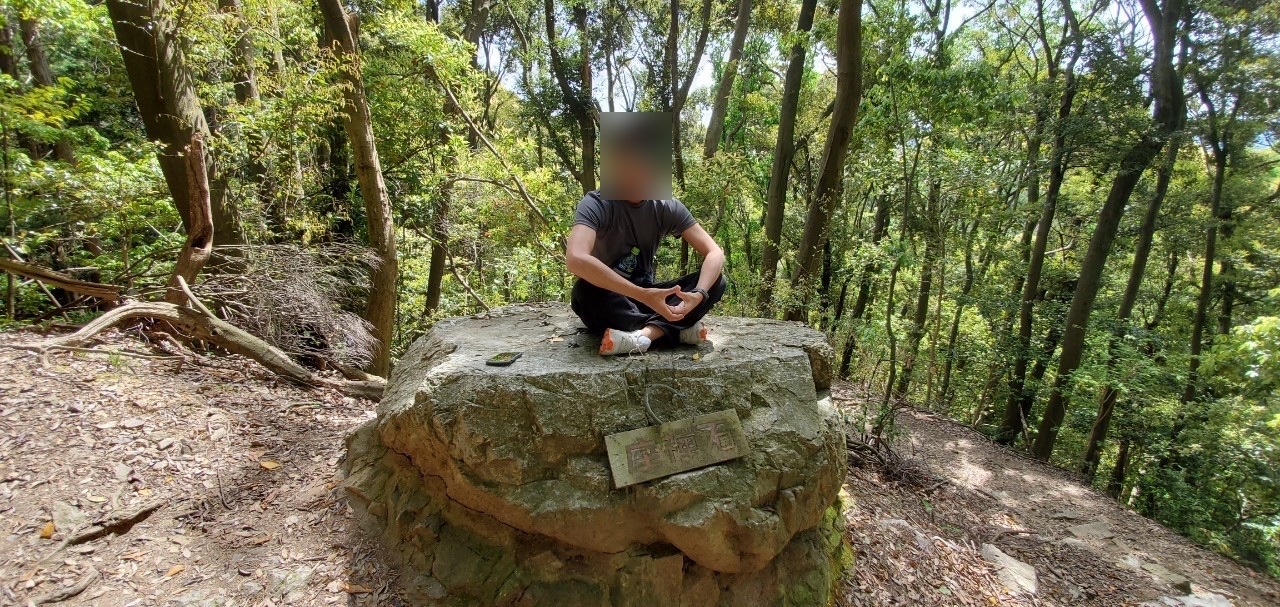 登山道の脇にある平らな大きな岩の上に座っている人。まわりは新緑の林