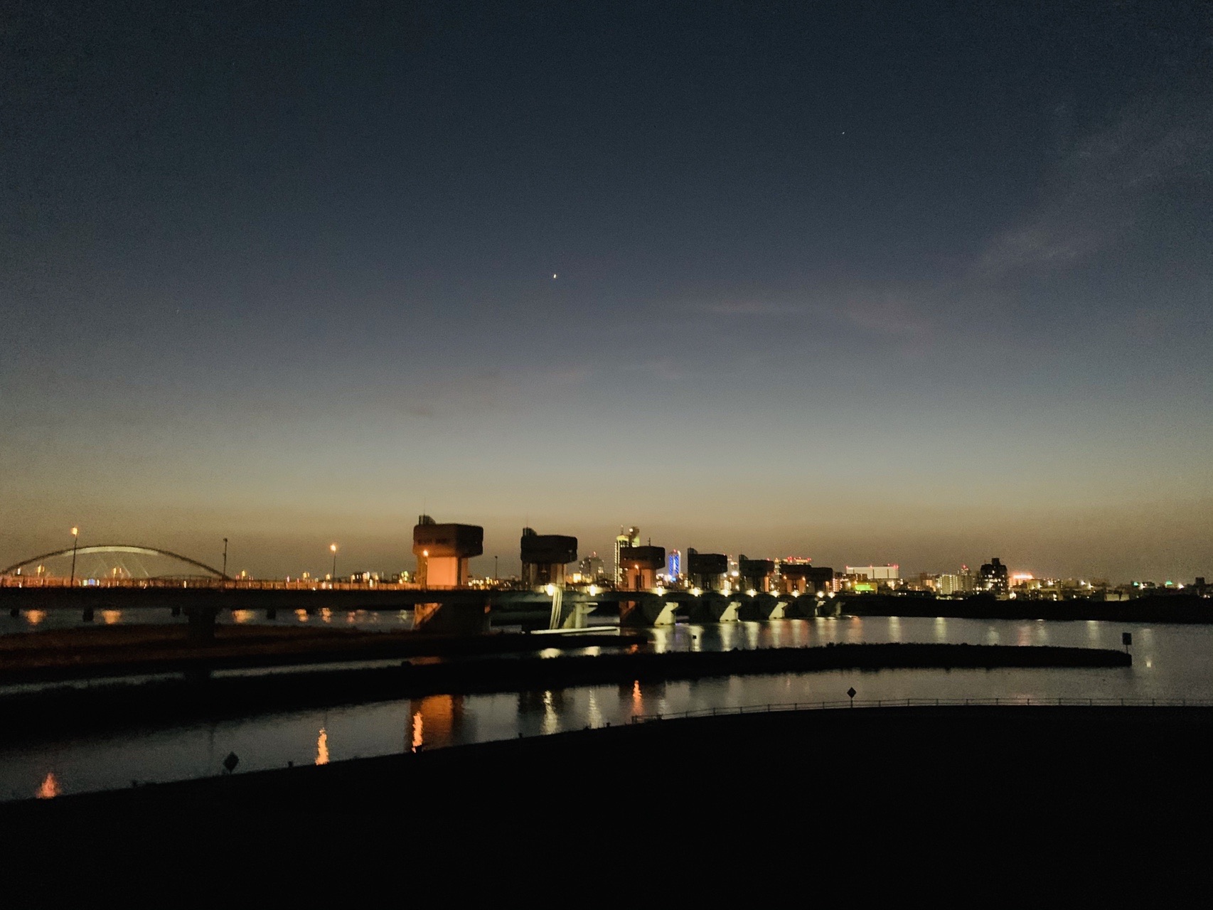 夜の川面に対岸の街明かりが長く伸びて映っている。奥に橋のシルエット